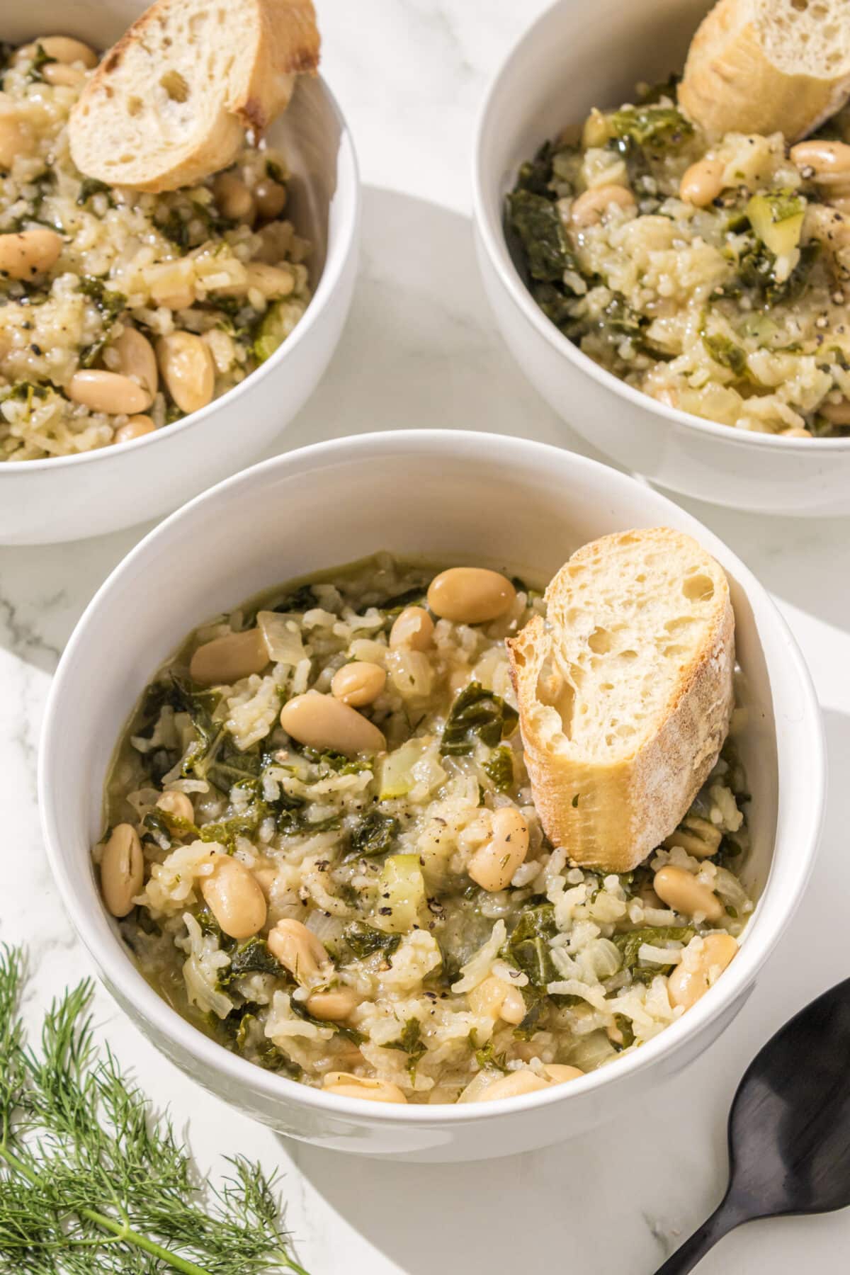 Three bowls of greens, rice, and bean soup with a piece of bread in each. Beside the bowls on the counter is a black metal spoon and a sprig of fresh dill.
