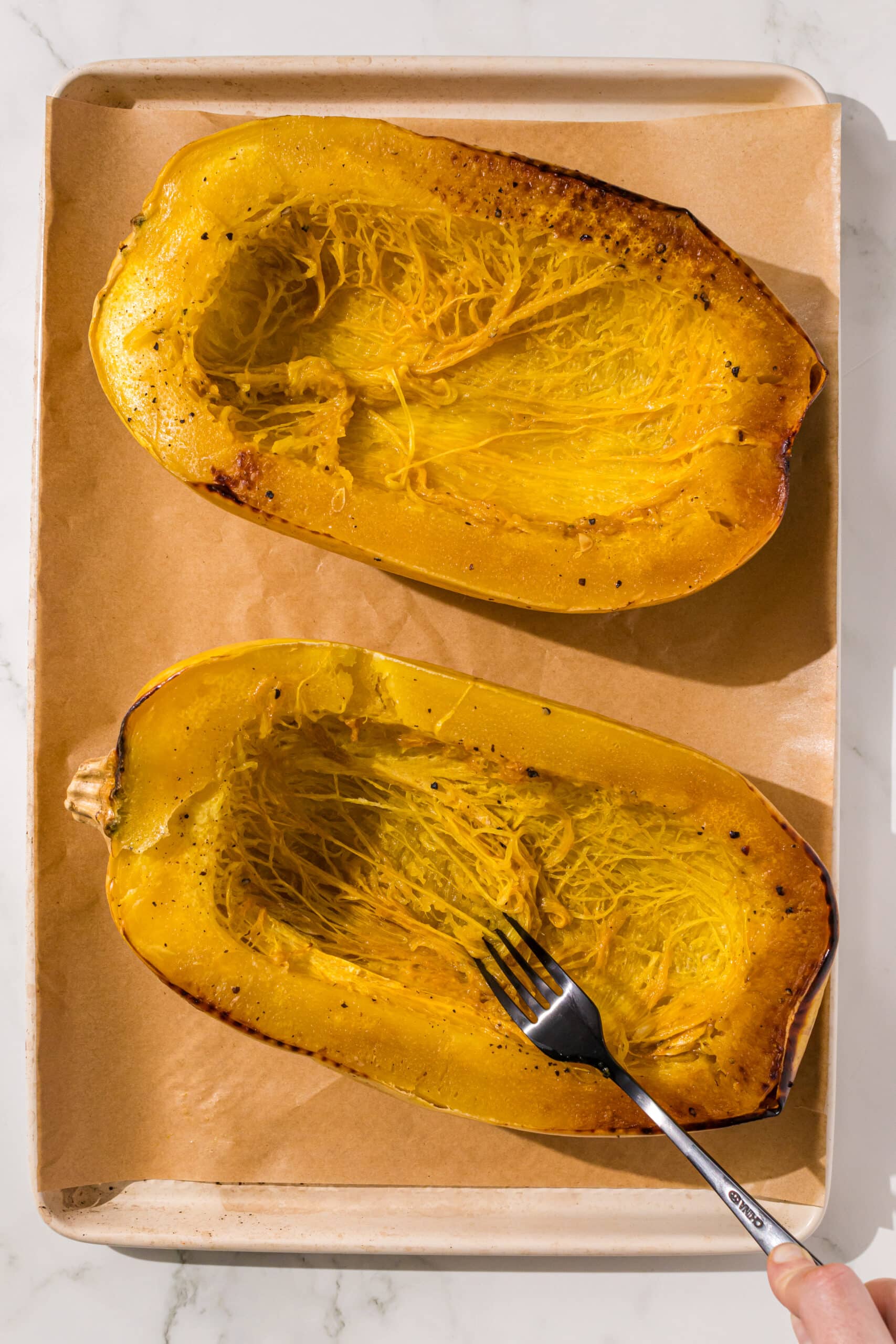 Two spaghetti squash halves with seeds scooped out and a black fork in the left one, sitting on a parchment lined baking sheet.