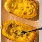 Two spaghetti squash halves on a baking sheet lined with parchment paper and a black metal fork in the left one.