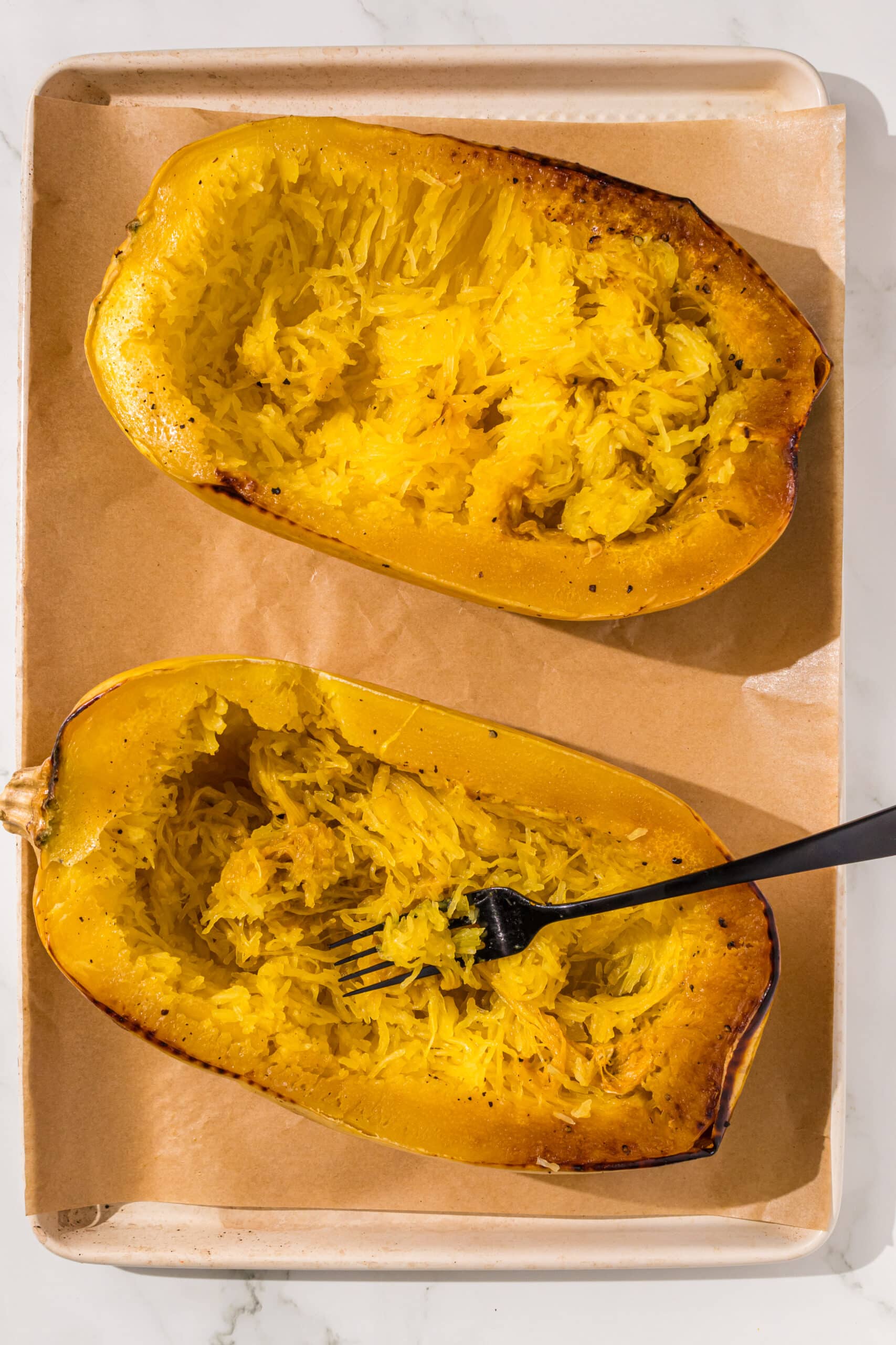 Two spaghetti squash halves on a baking sheet lined with parchment paper and a black metal fork in the left one.