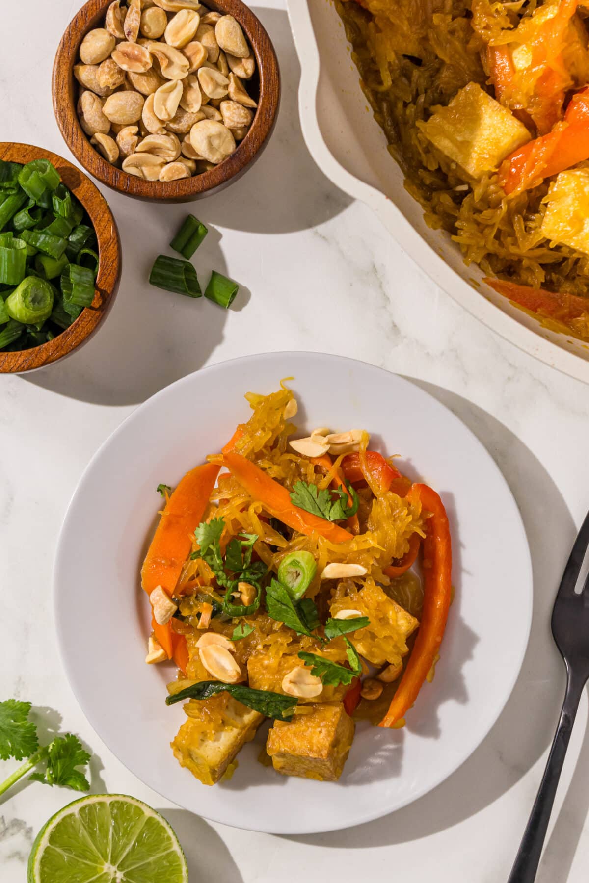 A small white plate with a serving of spaghetti squash stir fry on it, two small wooden bowls of peanuts and green onion, half of a lime, and a pot with the remaining stir fry in it.