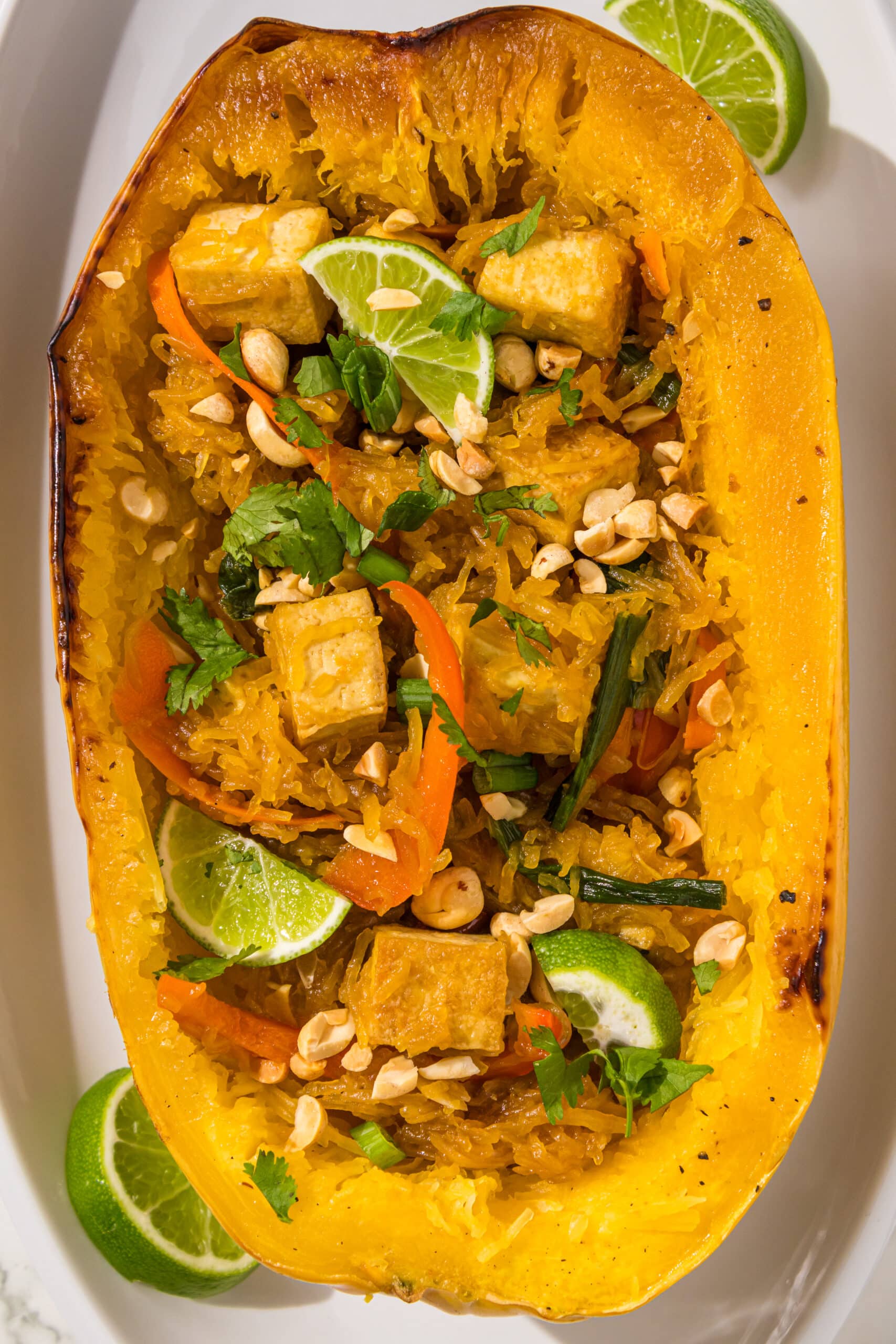 Half of a spaghetti squash filled with spaghetti squash tossed with tofu cubes, bell peppers, and lime wedges on top.