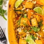 Half of a spaghetti squash filled with spaghetti squash, tofu, bell pepper slices, carrot slices, and lime wedges. Beside the squash is a black metal fork and a sprig of fresh cilantro laying on the counter.