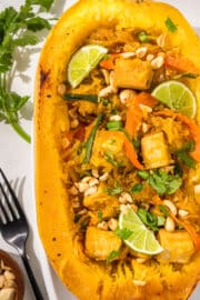 Half of a spaghetti squash filled with spaghetti squash, tofu, bell pepper slices, carrot slices, and lime wedges. Beside the squash is a black metal fork and a sprig of fresh cilantro laying on the counter.