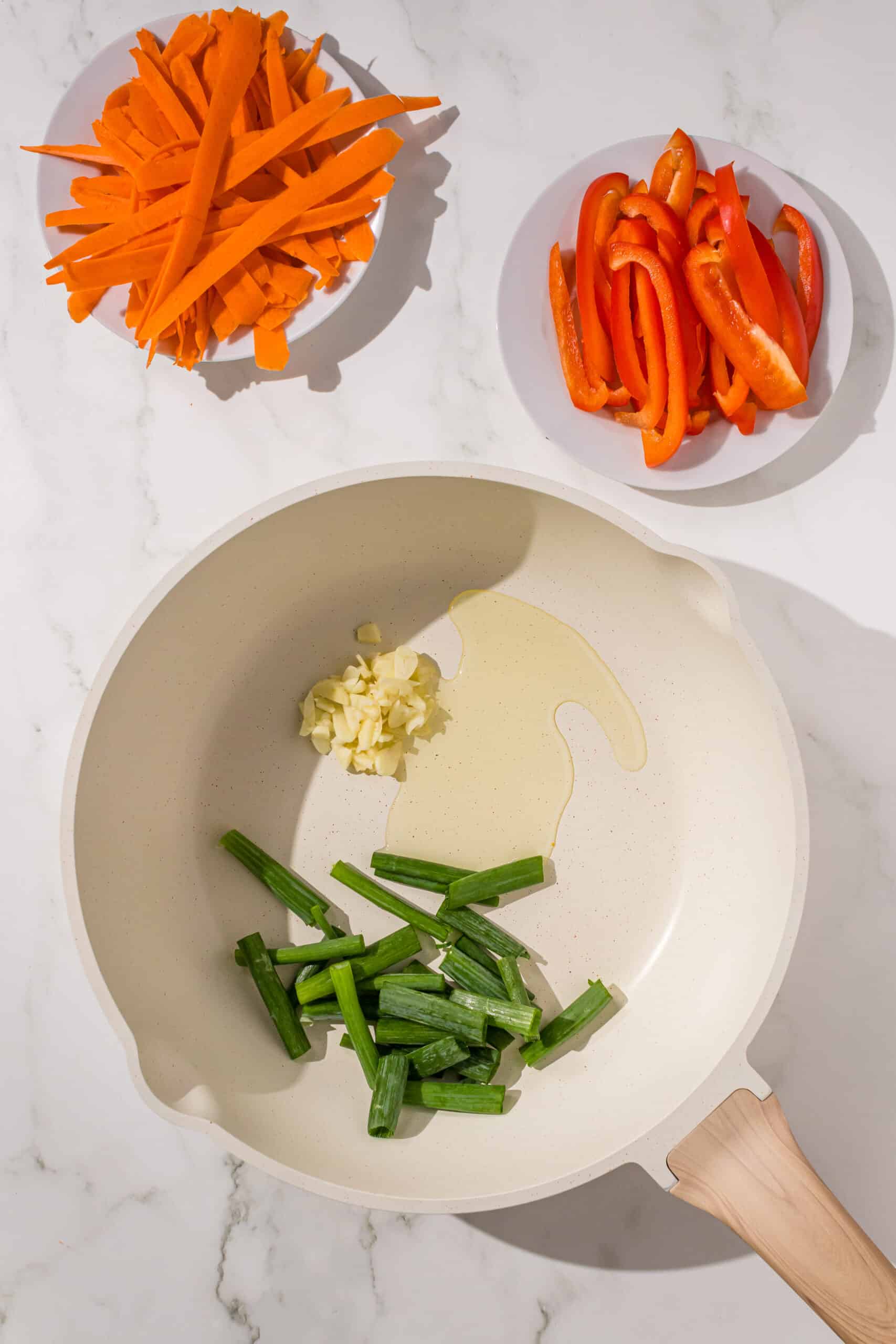 A small frying pan with scallions, garlic, and oil in it. Beside the pan is two small plates with shredded carrots and bell pepper on them.
