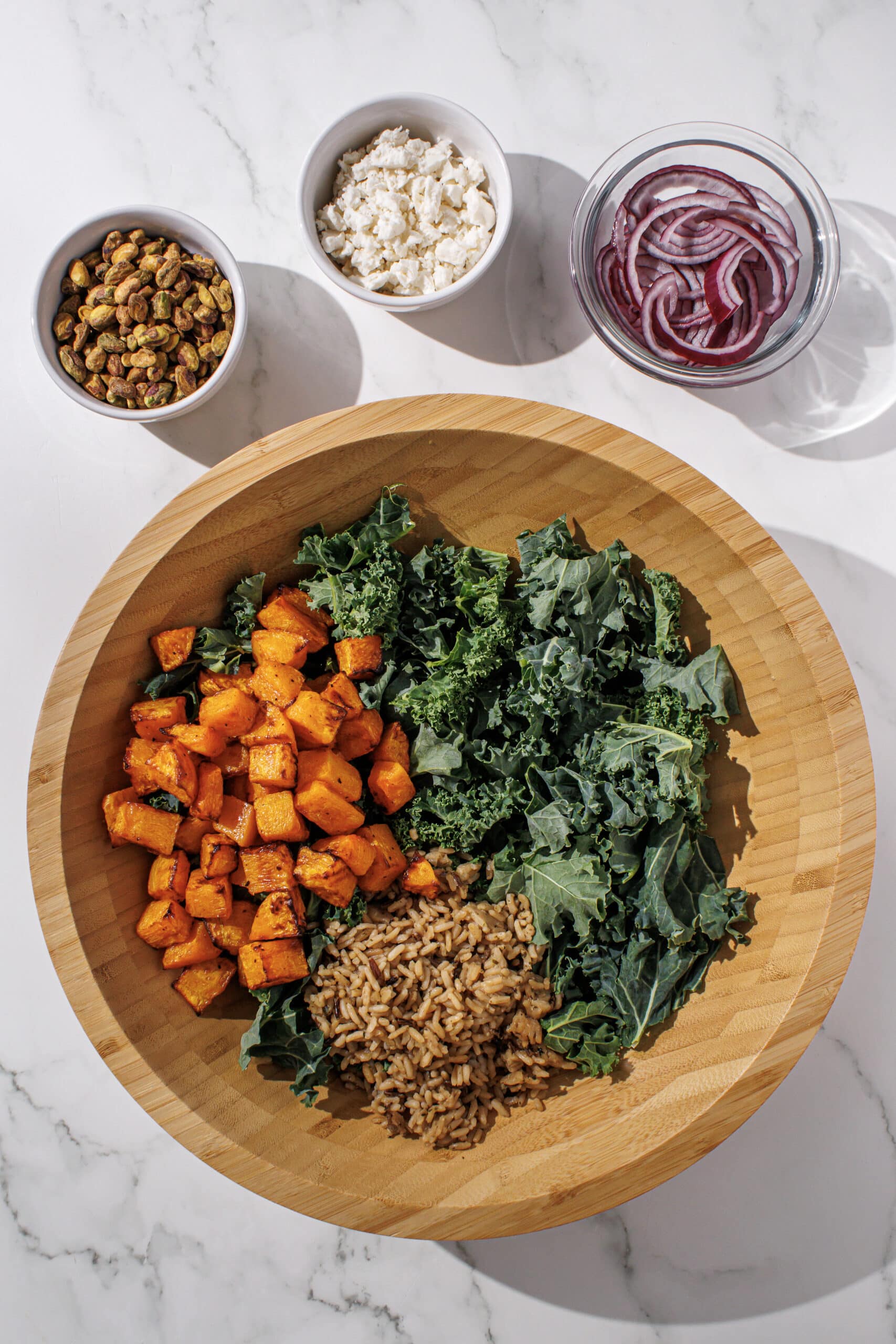 A large wooden bowl filled with kale and a pile of butternut squash and wild rice. Beside the bowl is three small bowls of pistachios, feta, and red onion sliced.