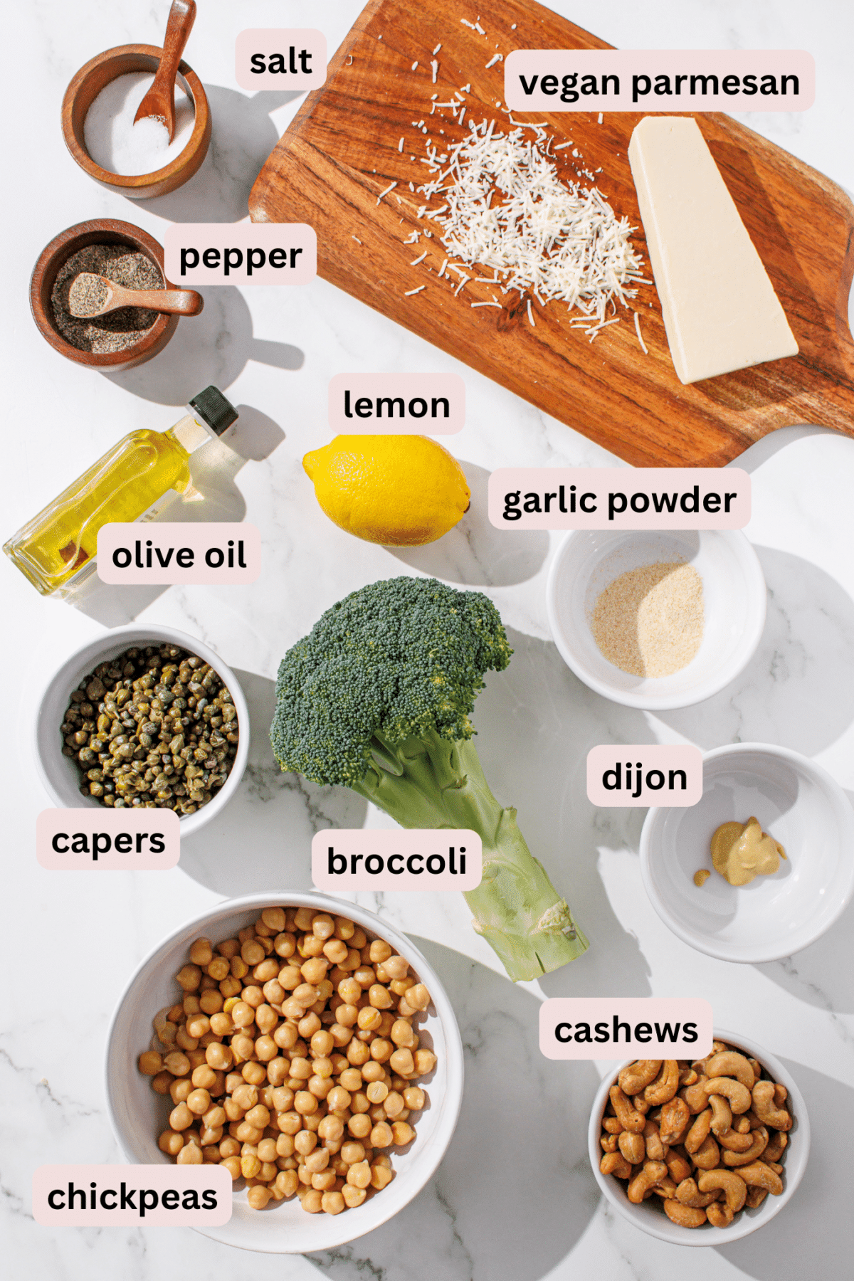 A photo of all ingredients for the broccoli caesar recipe laid out on a marble counter including: vegan parmesan, salt, pepper, lemon, olive oil, garlic powder, capers, broccoli, dijon, chickpeas, and cashews.