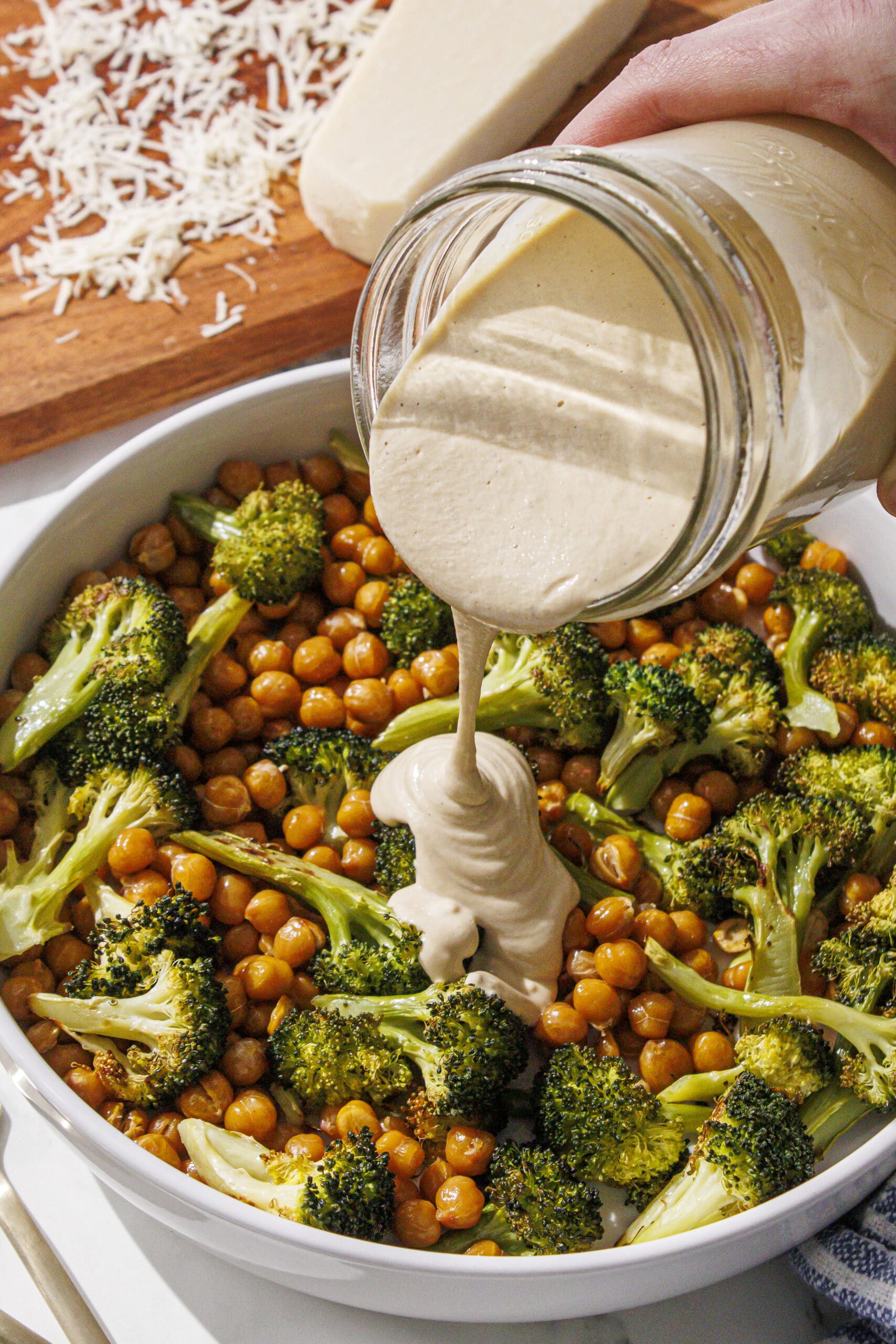 A white bowl filled with chickpeas and broccoli. There is a white hand coming from the right side holding a glass jar and pouring caesar dressing in the center. In the back of the photo is a brown cutting board with parmesan cheese on it.