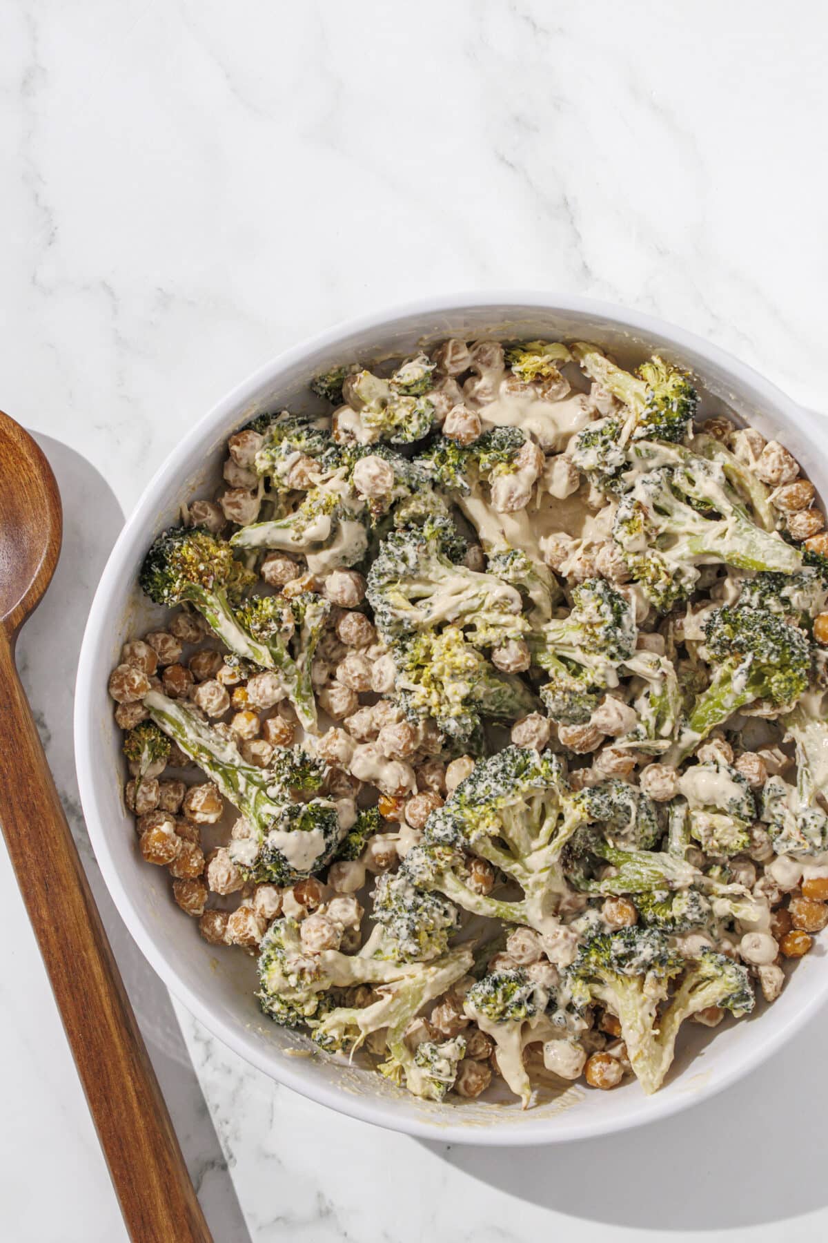 A white bowl filled with broccoli caesar salad. To the left of the bowl is a large wooden spoon.