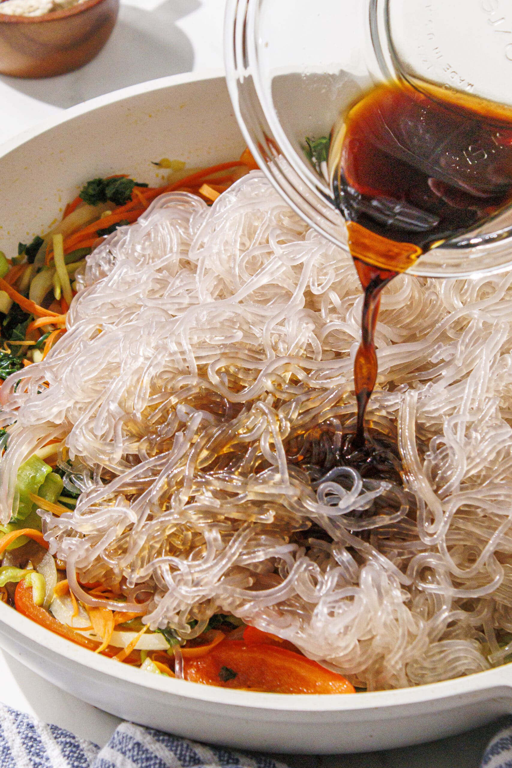 A white pan filled with sauteed veggies and sweet potato noodles with a clear bowl over top of soy sauce being poured in.