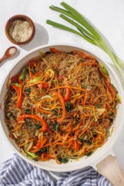 A large white bowl filled with Sweet Potato Noodle Japchae. Around the bowl is a blue and white striped towel, green onions, and sesame seeds in a mini wooden bowl.