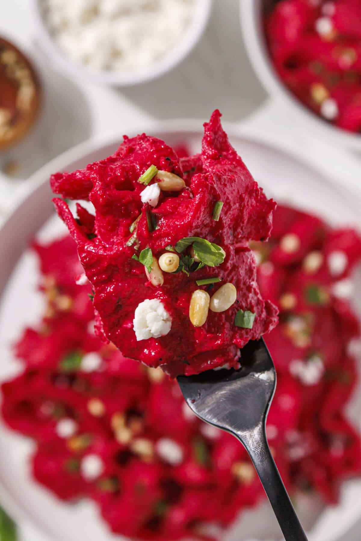 A fork with pink pasta on it over a plate with remaining pink pasta topped with pine nuts, feta cheese, and parsley. Plus, three small bowls of cheese, pine nuts, and more pasta.