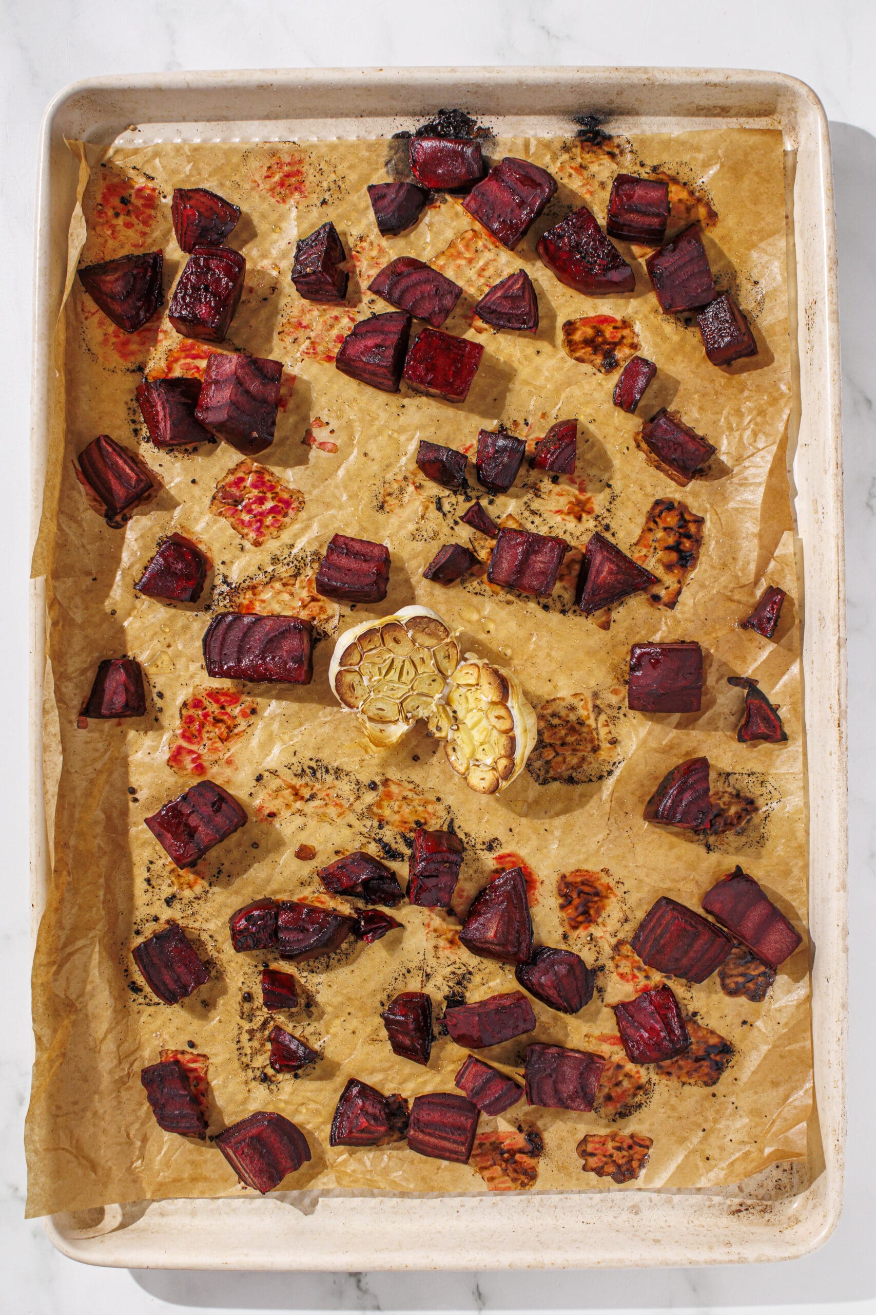 A baking pan lined with parchment paper filled with roasted beet cubes and a roasted garlic head.