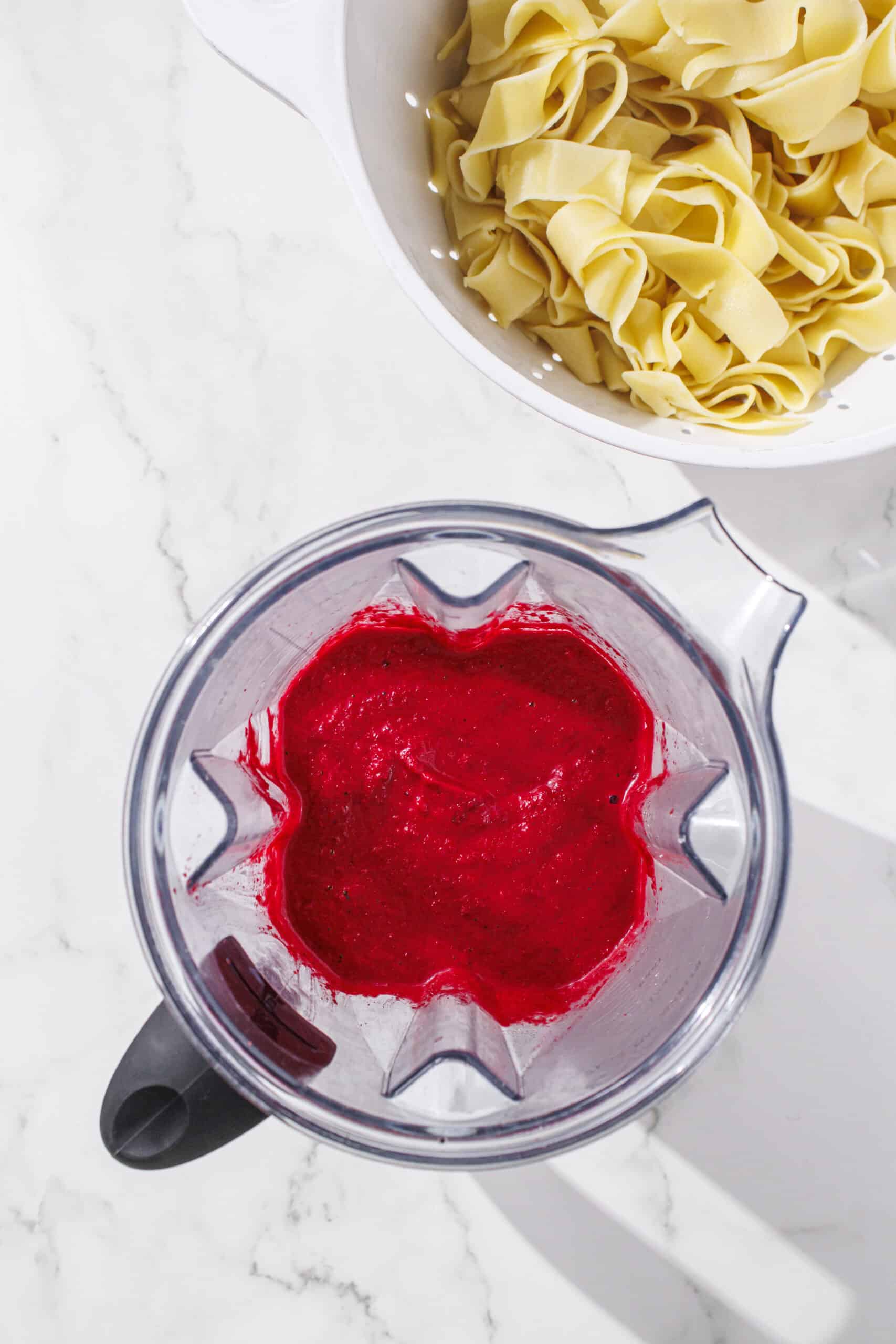 A blender filled with pink pasta sauce and a bowl of cooked pasta noodles.