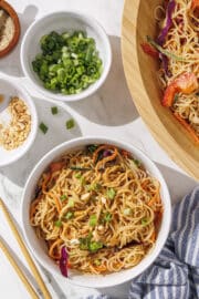 Spring Roll in a Bowl in a small white bowl with chopsticks, a blue towel, a bowl of green onion, a bowl of peanuts, and then a large wooden bowl with the remaining spring roll in a bowl in it.