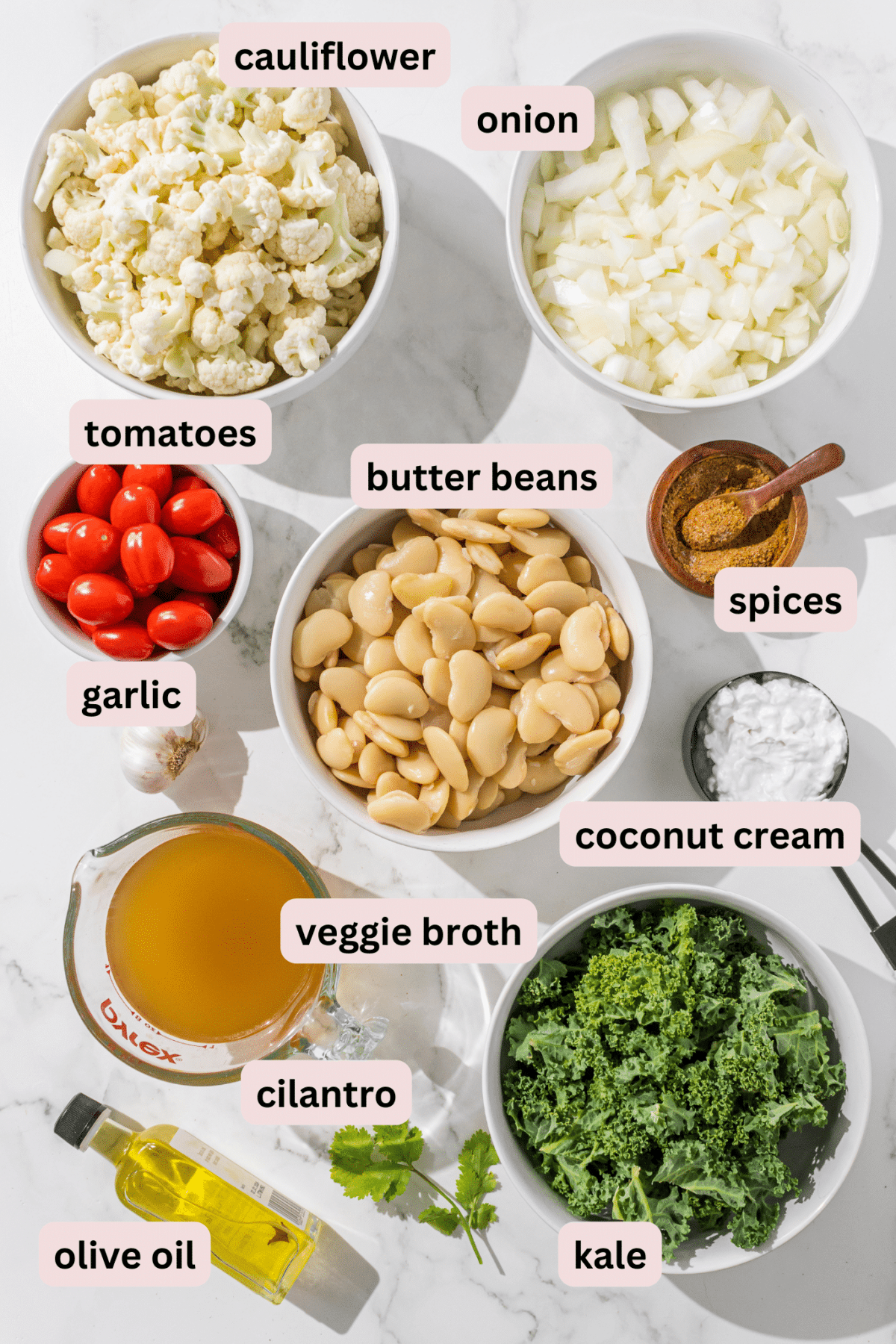 Ingredients laid out in bowls on a marble counter and labeled including: cauliflower, diced onion, tomatoes, butter beans, spices, coconut cream, kale, veggie broth, cilantro, fresh garlic, and olive oil.