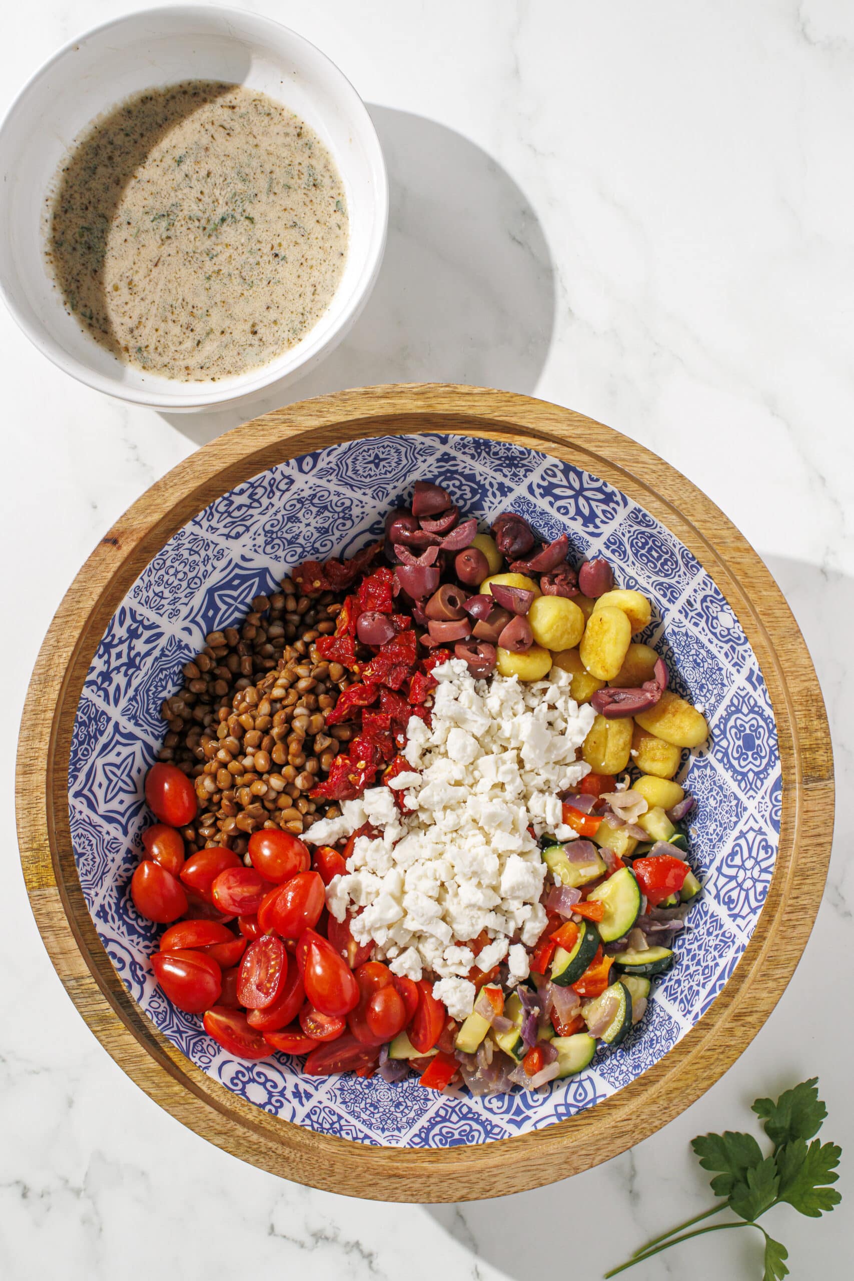 A brown and blue bowl filled with gnocchi, feta, tomatoes, zucchini, red onion, bell pepper, and lentils. Next to the bowl is a sprig of parsley and a small white bowl filled with dressing.