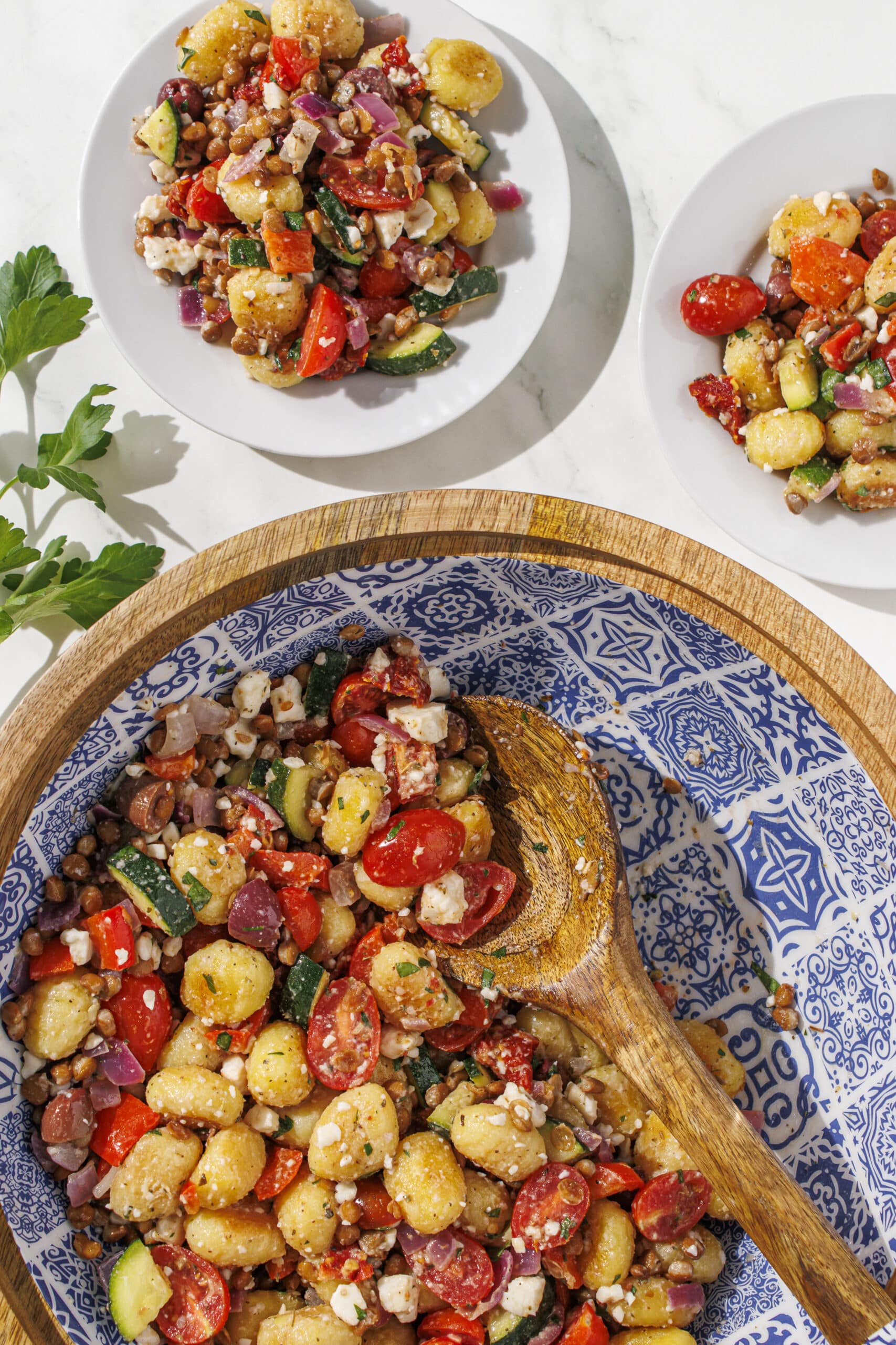 A brown and blue bowl filled with mediterranean gnocchi and a brown wooden spoon. Above the bowl is two small white plates with a serving of gnocchi on them and a sprig of parsley.
