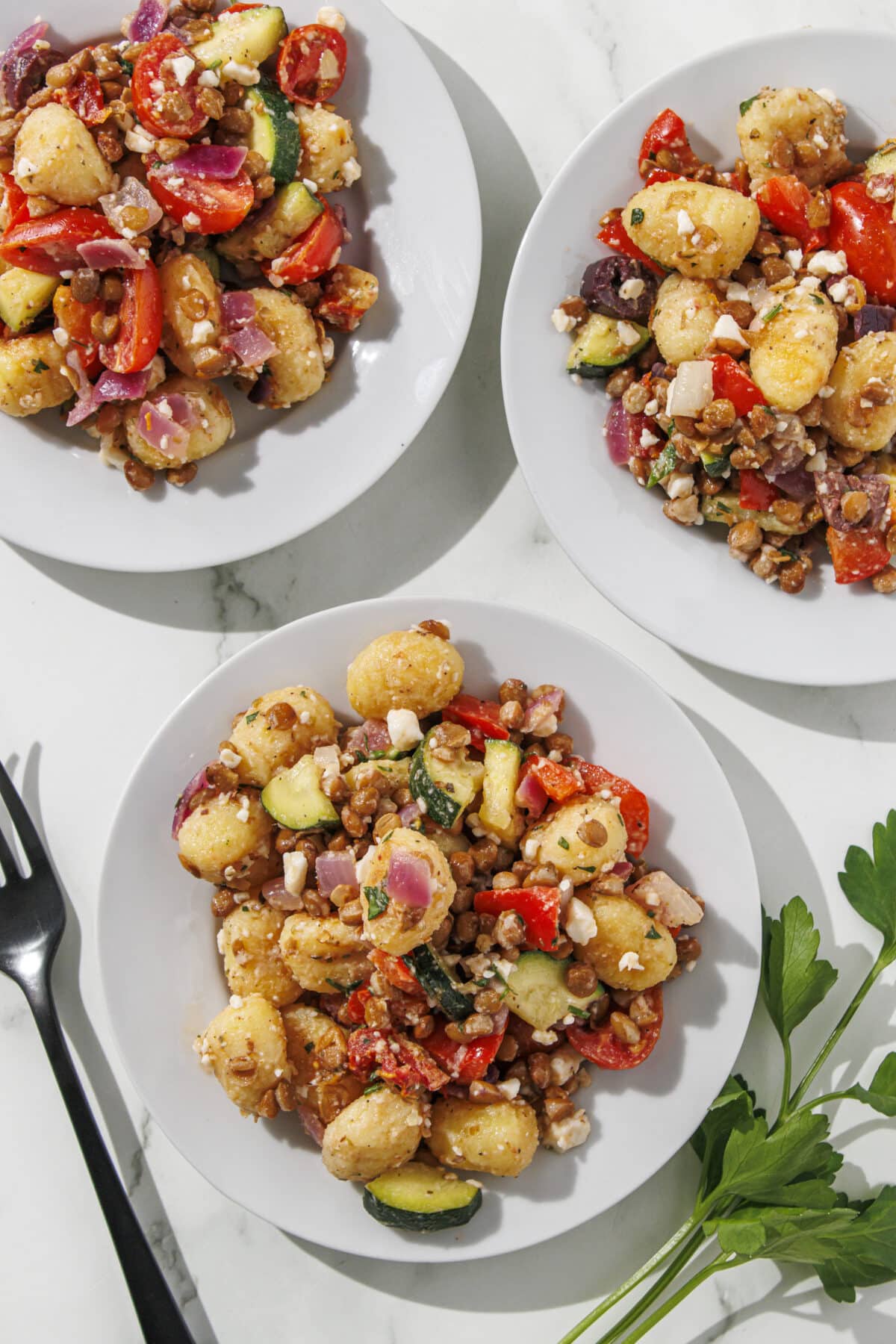 Three white plates each with a serving of mediterranean gnocchi on them. To the left of the plates is one black fork and to the right is fresh parsley.