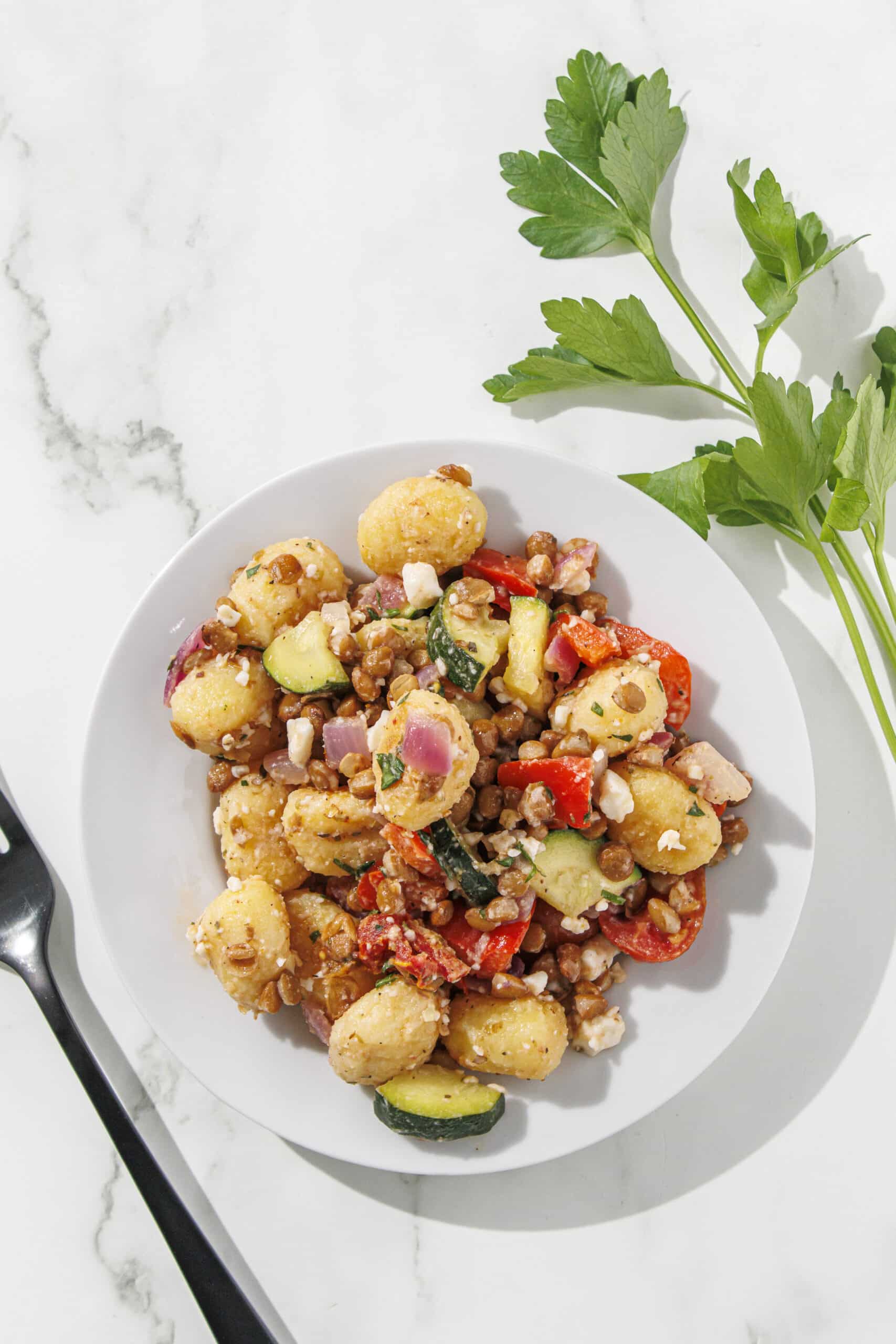 A small white plate filled with mediterranean gnocchi. Beside the plate is a sprig of parsley and a black fork.