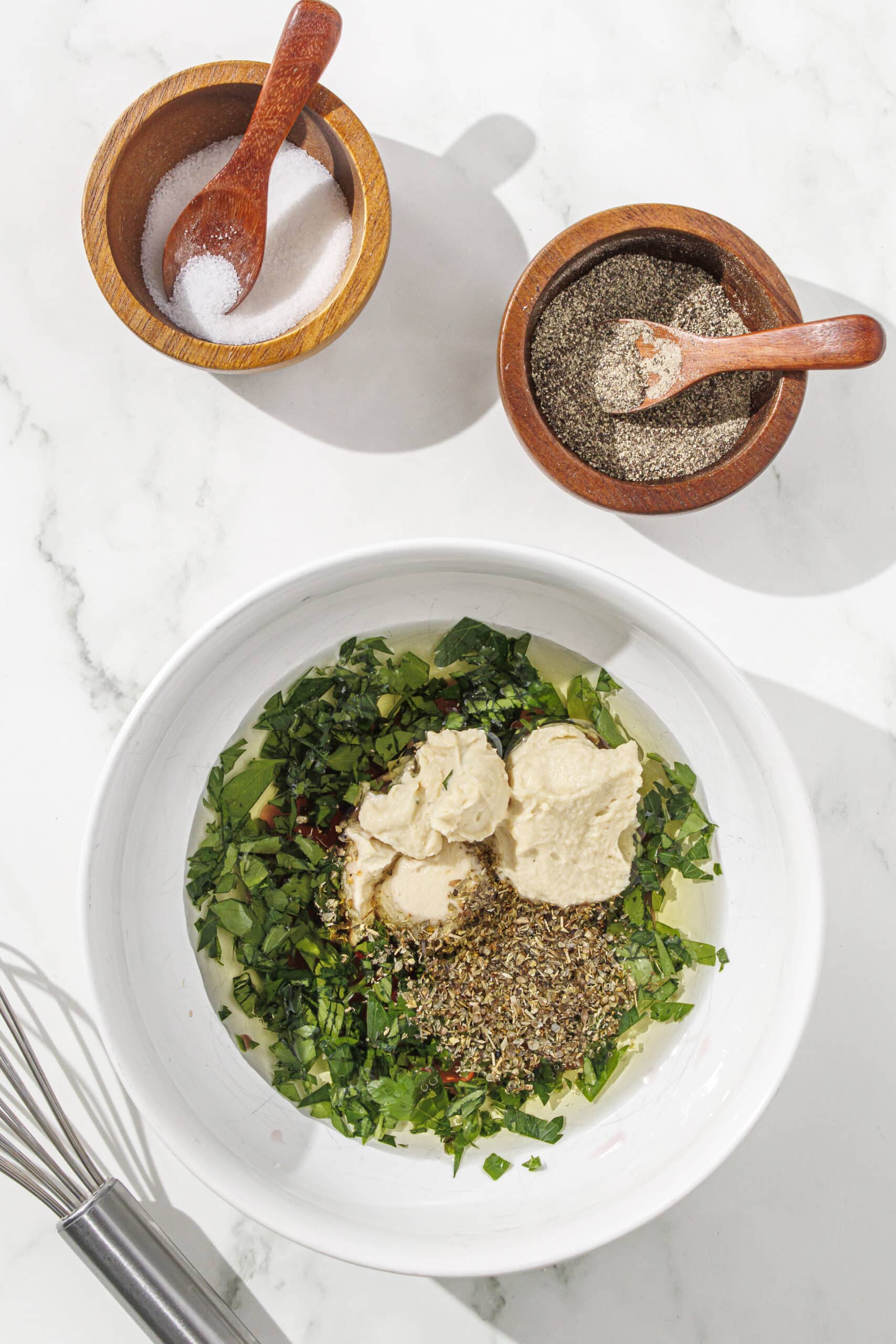 A white bowl filled with hummus, olive oil, vinegar, and parsley. There is a silver whisk next to the bowl along with two wooden bowls filled with salt and pepper.