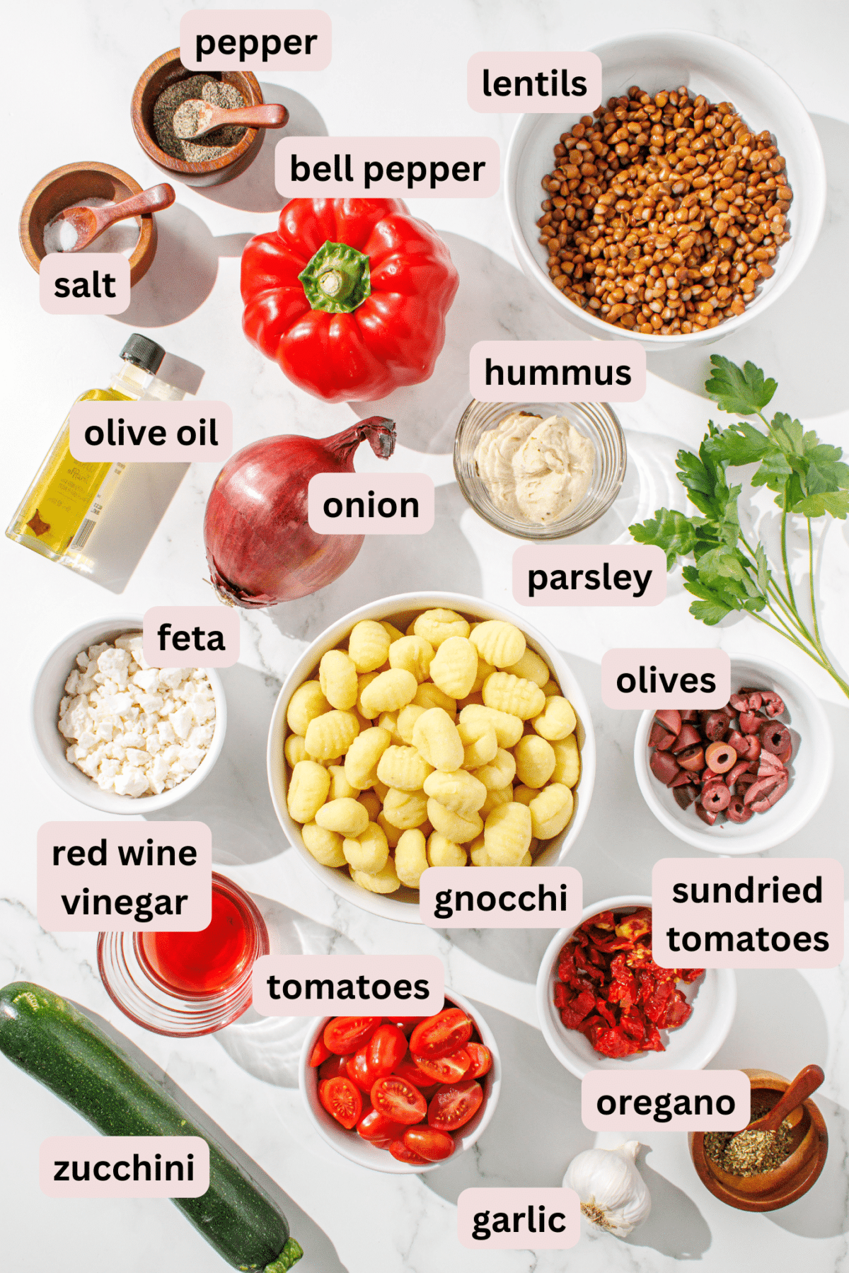 All of the ingredients required for mediterranean gnocchi laid out on a marble surface including: salt, pepper, lentils, bell pepper, olive oil, hummus, onion, parsley, feta, gnocchi, sundried tomatoes, olives, red wine vinegar, tomatoes, oregano, garlic, and zuchini.