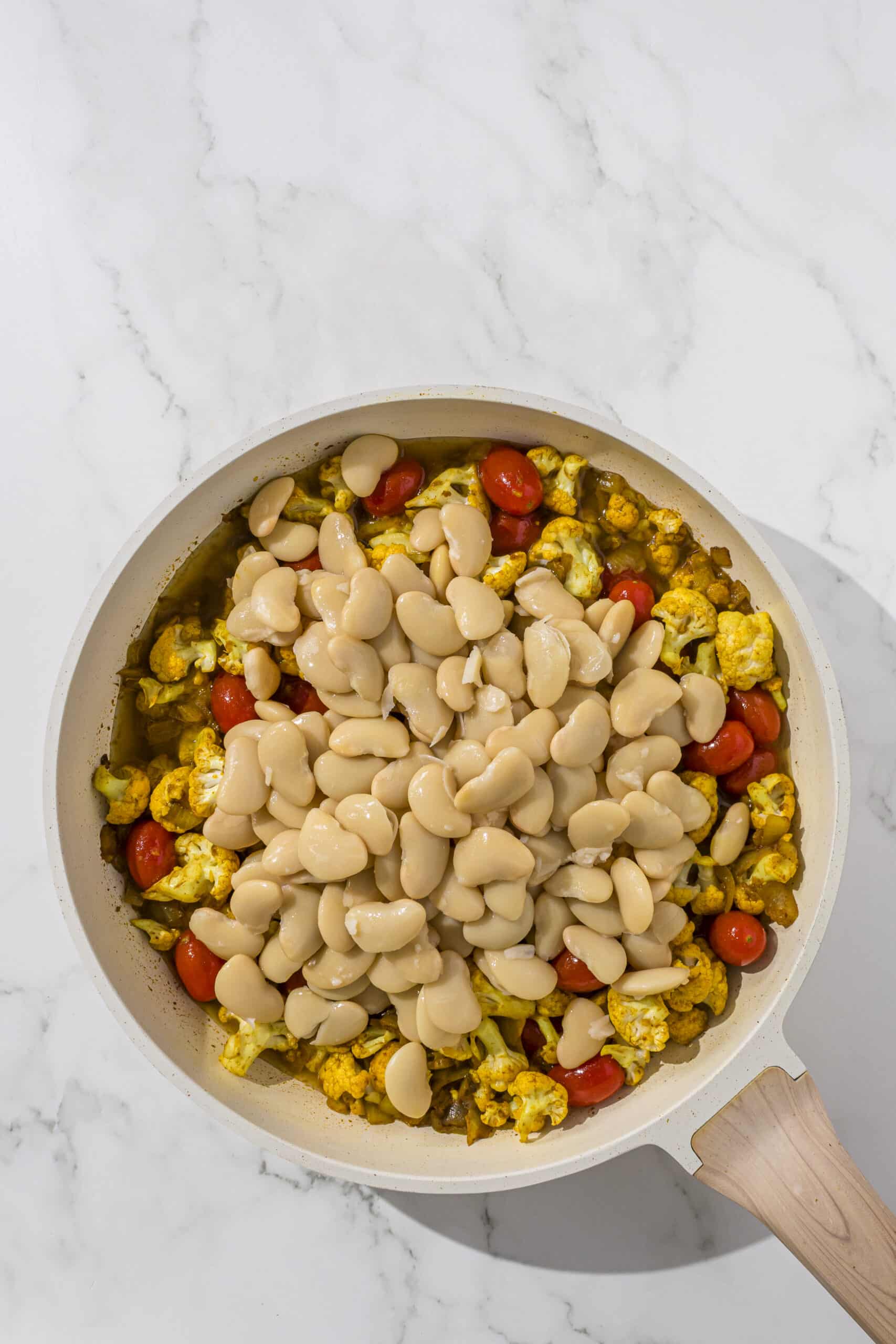 A large pan filled with chopped onion, cauliflower, cherry tomatoes, and butter beans.