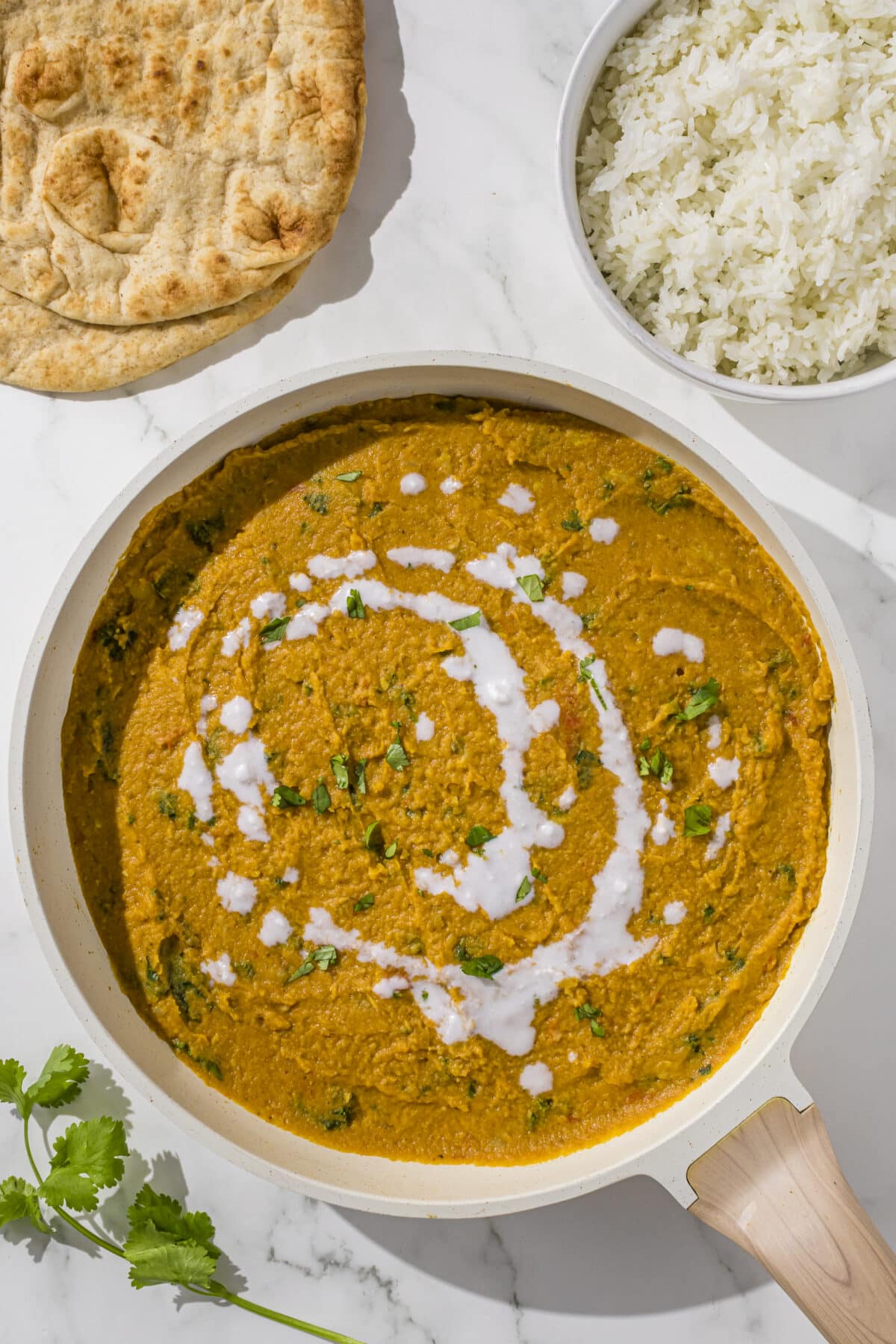 A large white pan filled with yellow curry. Beside the pan is two rounds of pita bread, a white bowl of white rice, and a sprig of fresh cilantro.