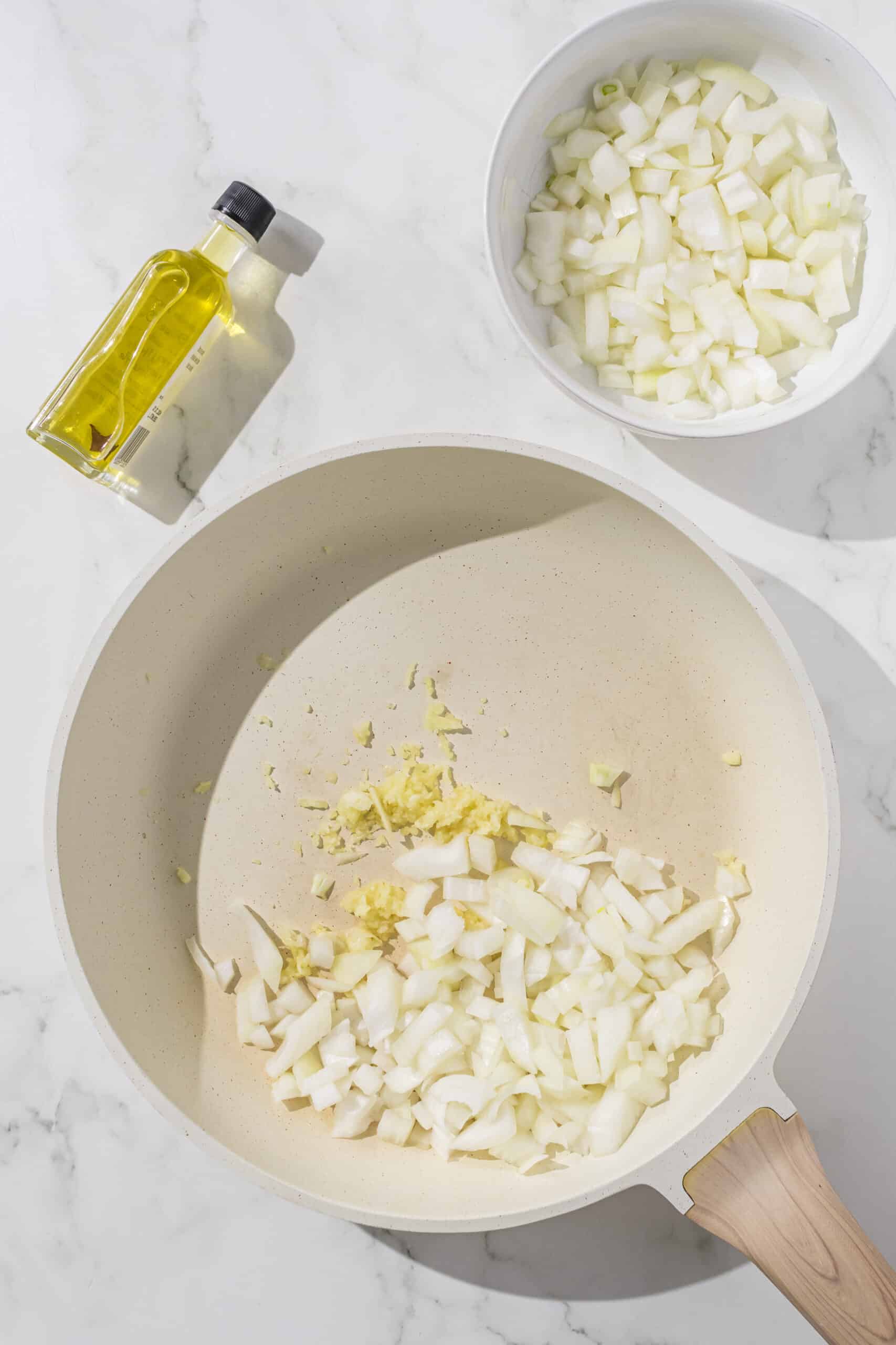 A large pan filled with fresh onion and garlic chopped. Beside the pan is a small bowl of chopped white onion and a small bottle of olive oil.