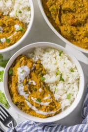 Two white bowls filled half with yellow curry and half with rice. Beside the bowls is two black metal forks, a blue and white striped towel, and a large white bowl filled with remaining yellow curry.