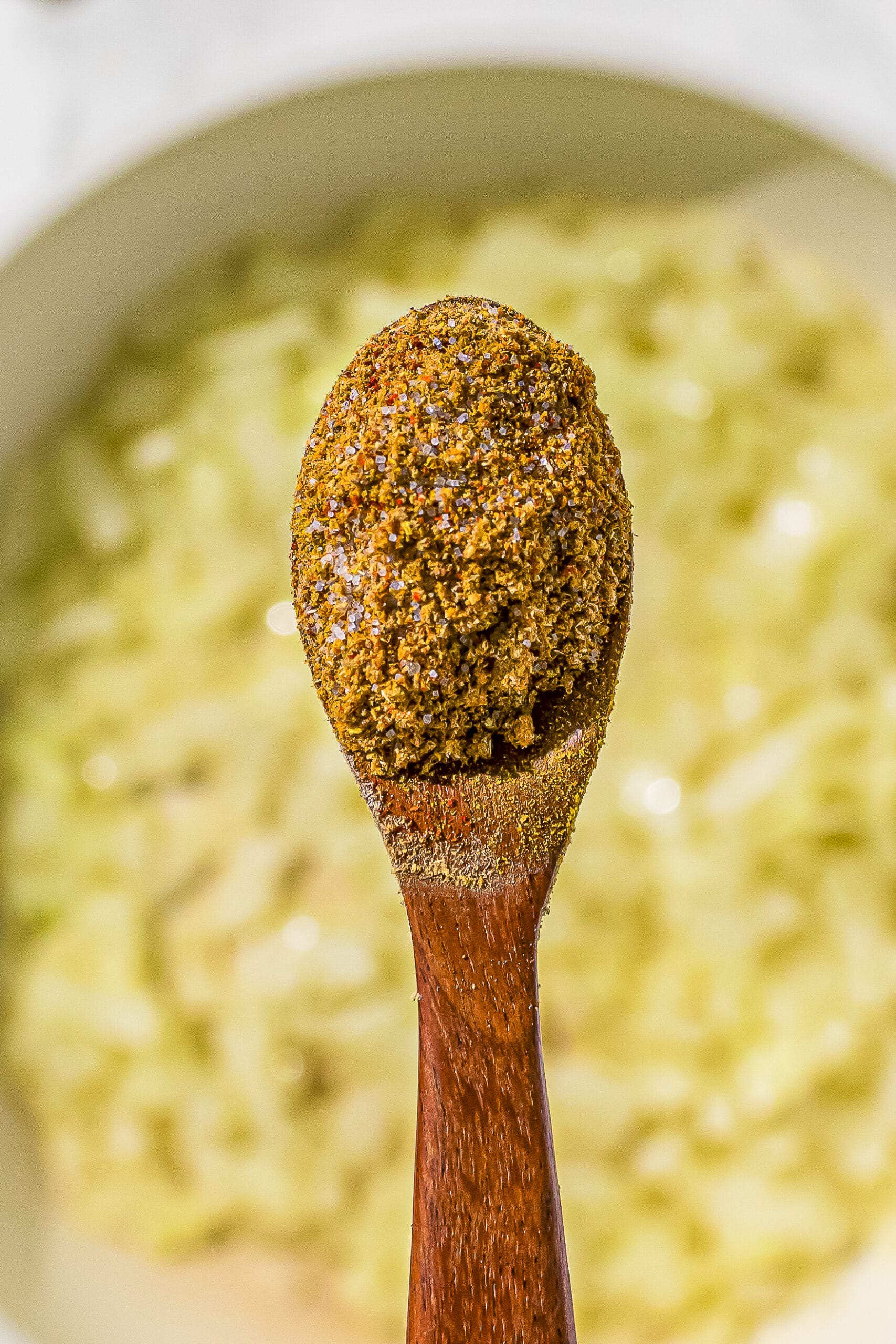 A pan in the background filled with onion being sauteed in oil. In the center of the image is a wooden spoon filled with spices.