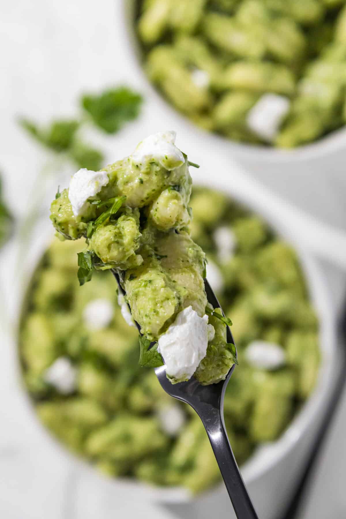 A bite of the all your greens pasta on a black fork being held overtop two large white bowls of the pasta. Next to the bowls is another black fork laying on the counter and a sprig of parsley.