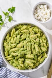 All Your Greens Pasta in a large white bowl. Around the bowl is a blue and white striped towel, a sprig of parsley, a small white bowl of vegan cheese, and a gold spoon.