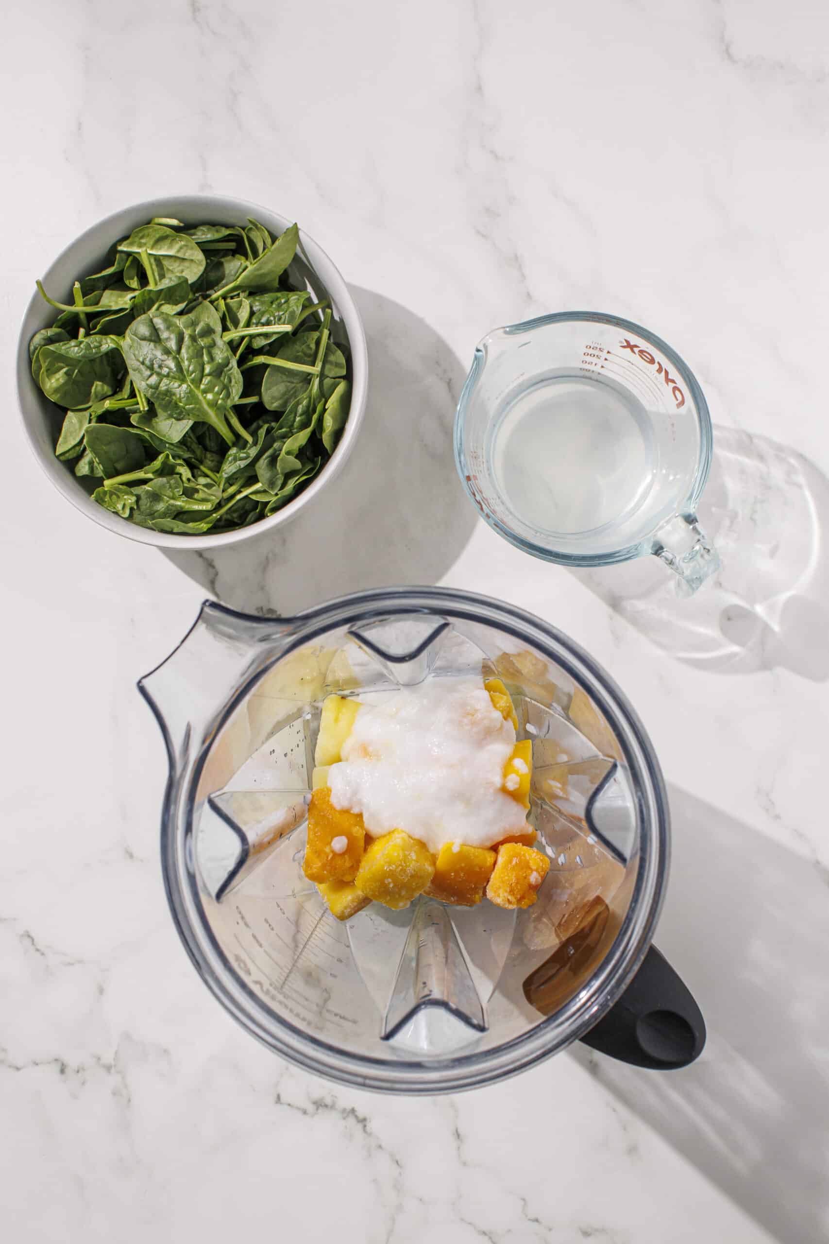 Frozen mango, frozen pineapple, and yogurt in a blender with a bowl of spinach and a cup of water beside it.