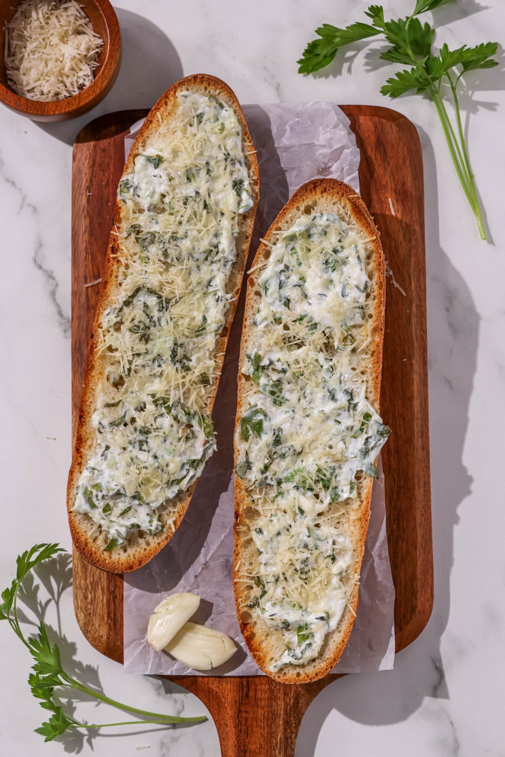 Two loaves of garlic bread on a wooden cutting board with a bowl of parmesan cheese and fresh herbs.