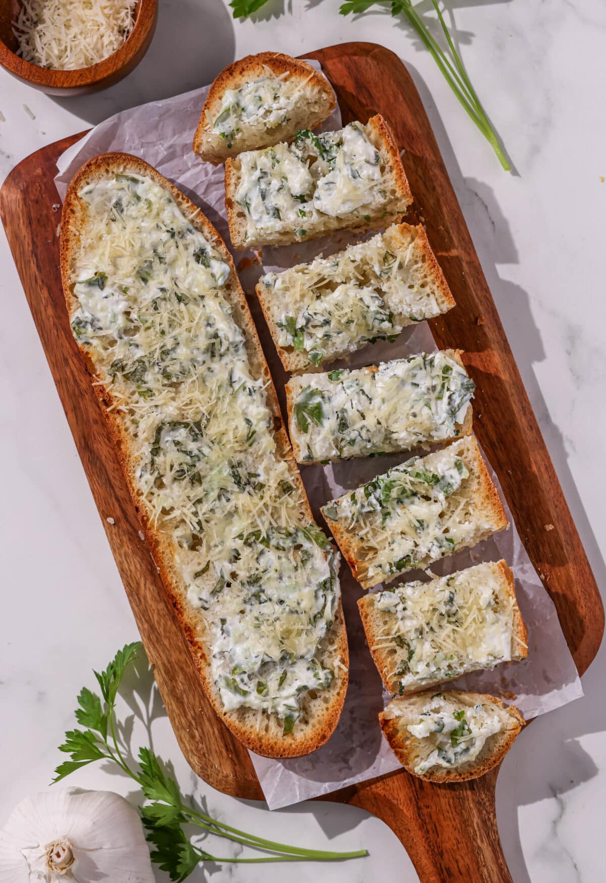 A wooden cutting board filled with half of a baguette and a second half of a sliced baguette garlic bread. Around the cutting board is fresh parsley and a small wooden bowl of parmesan.