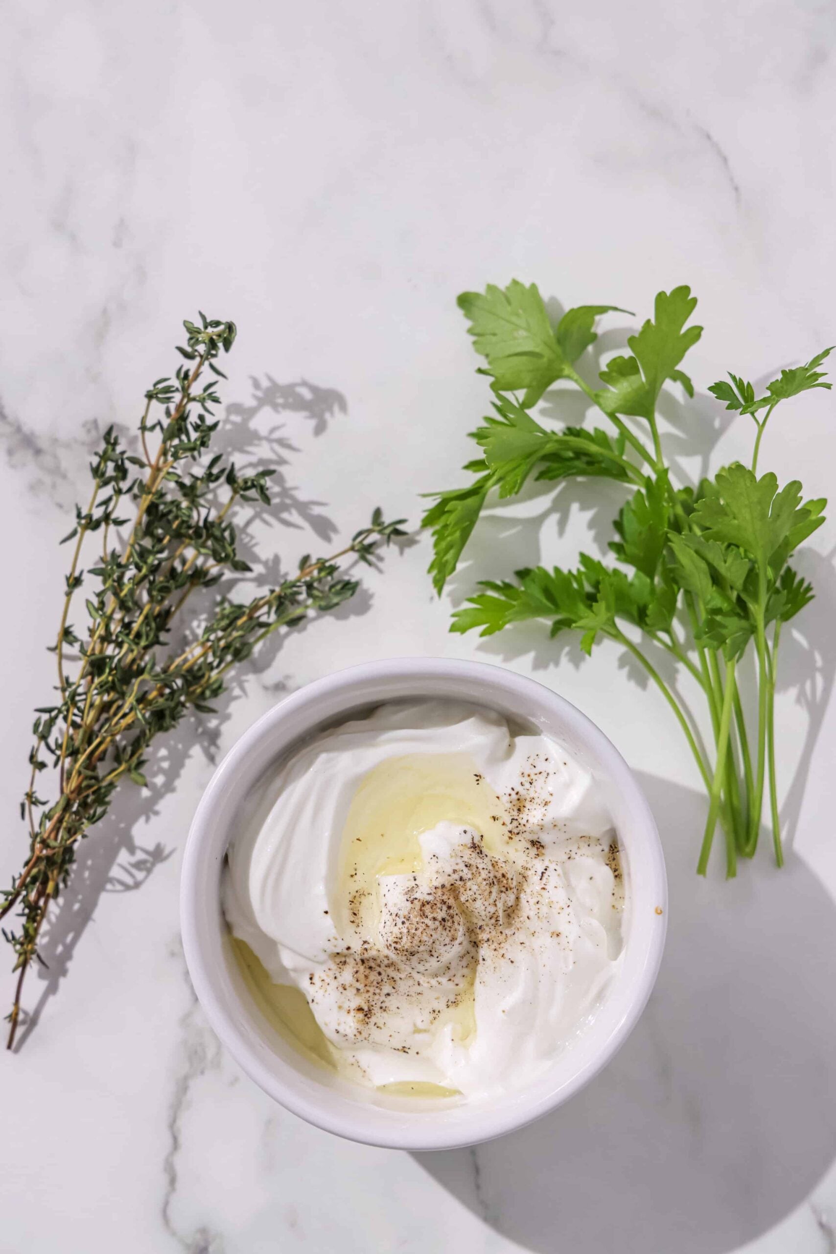 A small bowl of yogurt, oil, and black pepper. Around the bowl is fresh thyme and parsley.
