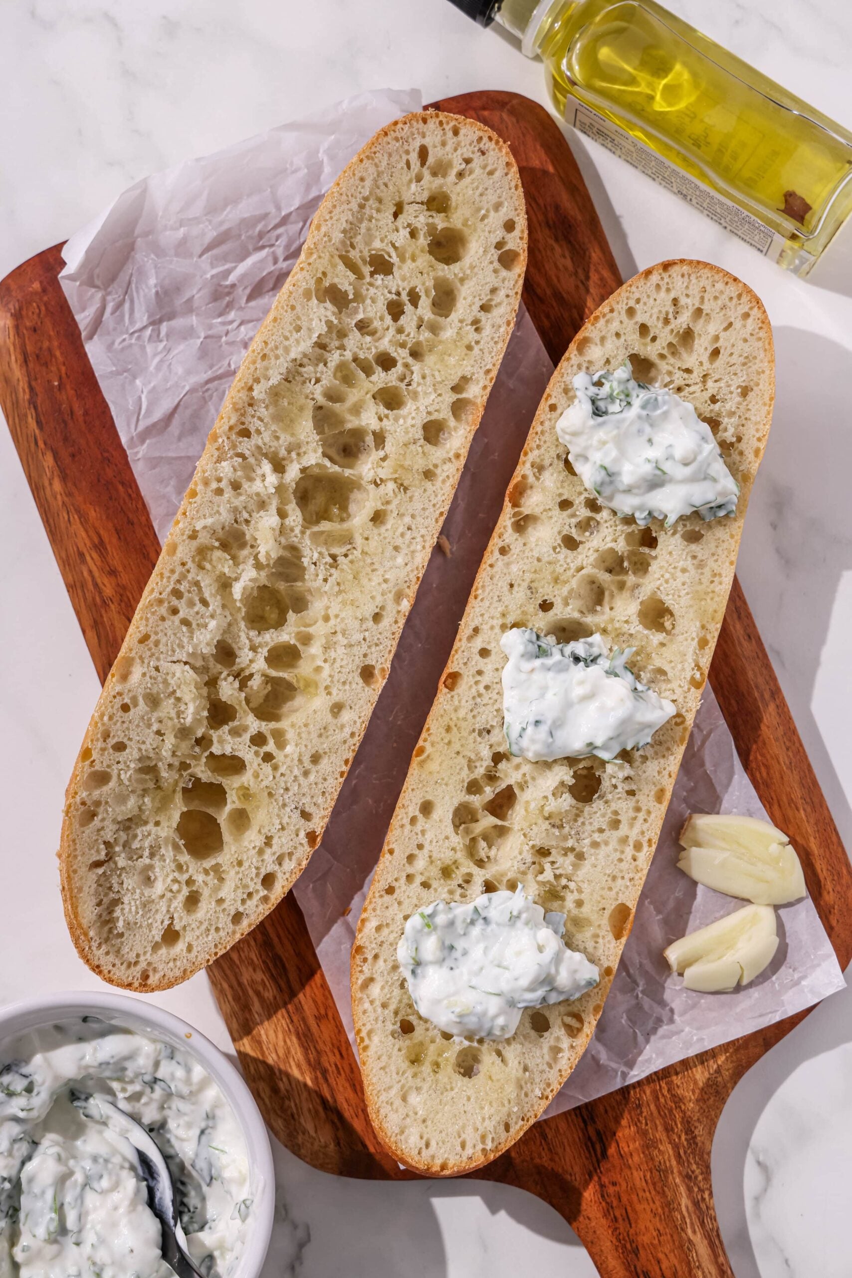 A baguette sliced in half on a wooden cutting board topped with dollops of cheese.
