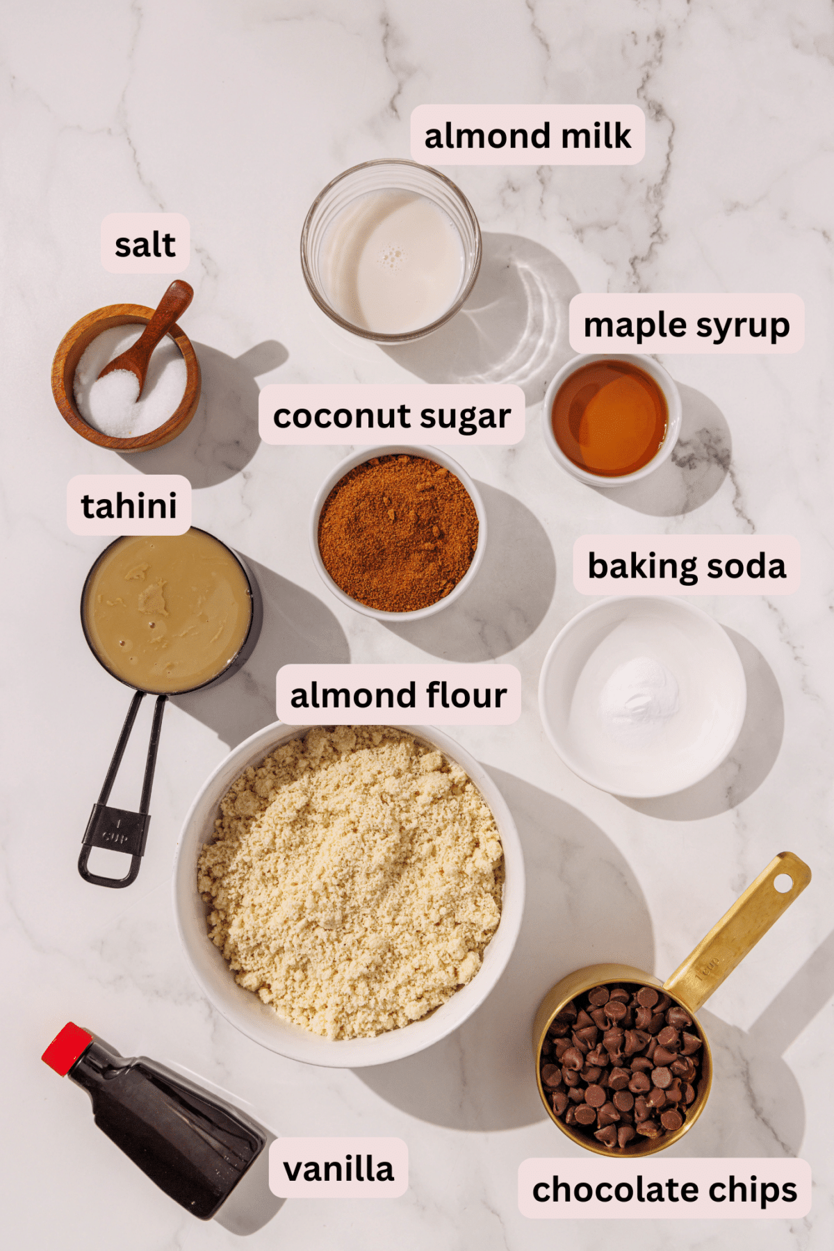 Ingredients needed to make vegan tahini blondies laid out on a marble counter and labeled including: almond milk, maple syrup, salt, coconut sugar, baking soda, tahini, almond flour, chocolate chips, and vanilla extract.