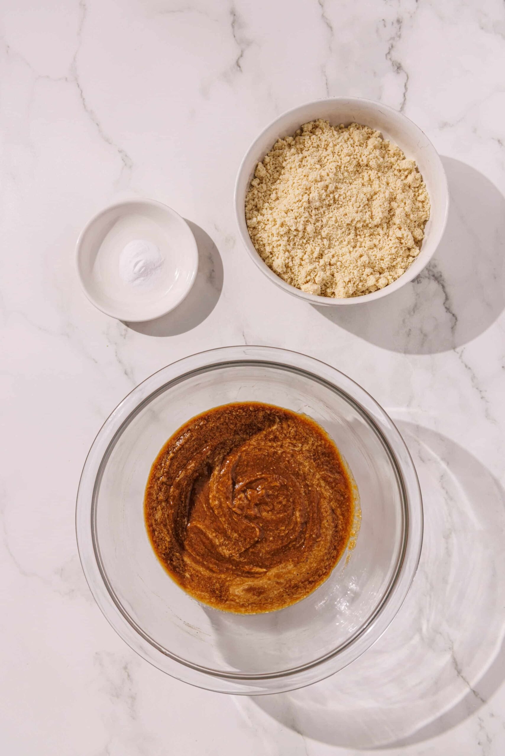 A glass bowl of vegan tahini blondie batter with two small white glass bowls above it of baking soda and almond flour.