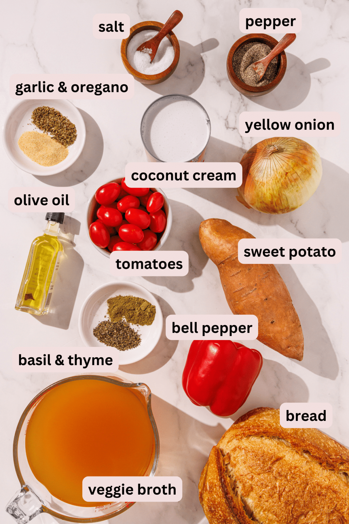 Ingredients laid out on a marble counter required to make glowing skin soup: sweet potato, veggie, broth, a loaf of bread, bell pepper, basil and thyme, tomatoes, onion, coconut cream, olive oil, garlic and oregano, salt, and pepper.