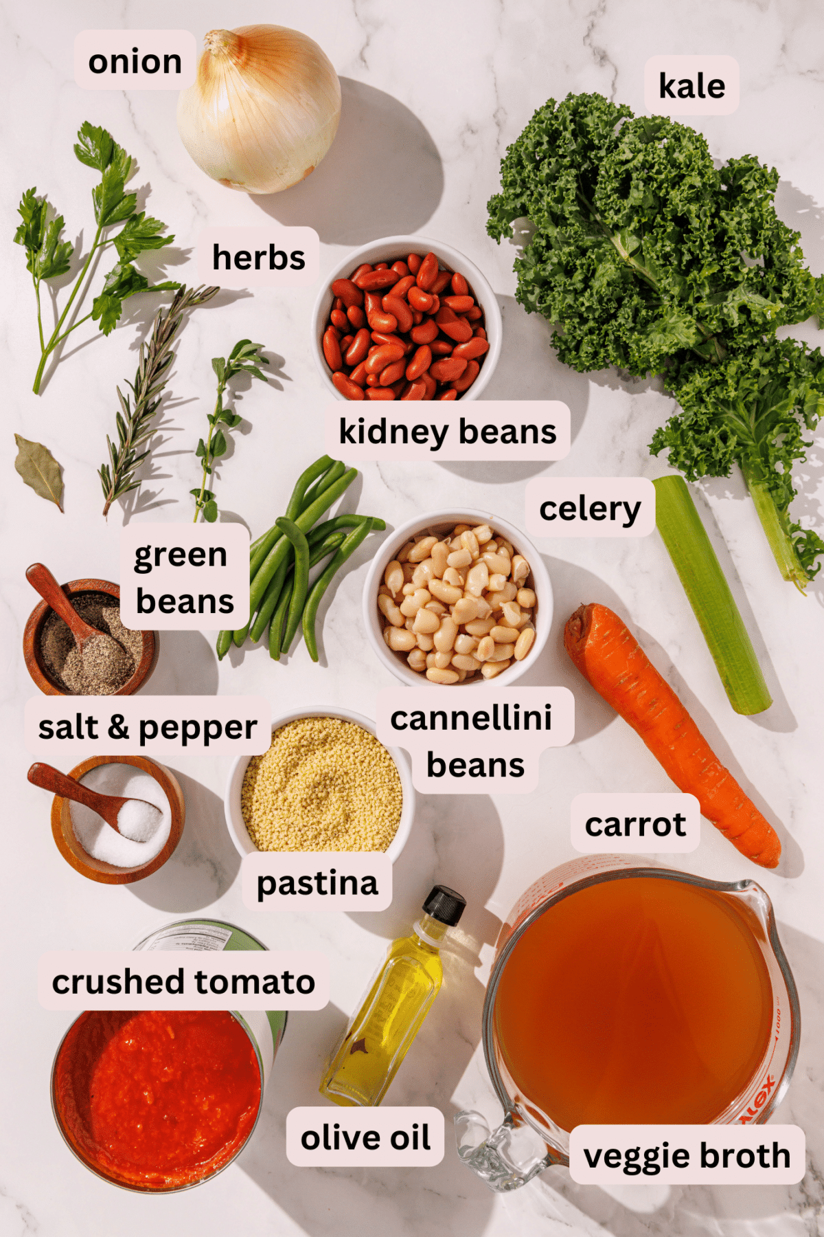 Ingredients needed to make mama's minestrone soup laid out on the counter and labeled including: onion, kale, kidney beans, celery, herbs, green beans, cannellini beans, carrot, pastina, salt and pepper, crushed tomato, veggie broth, and olive oil.