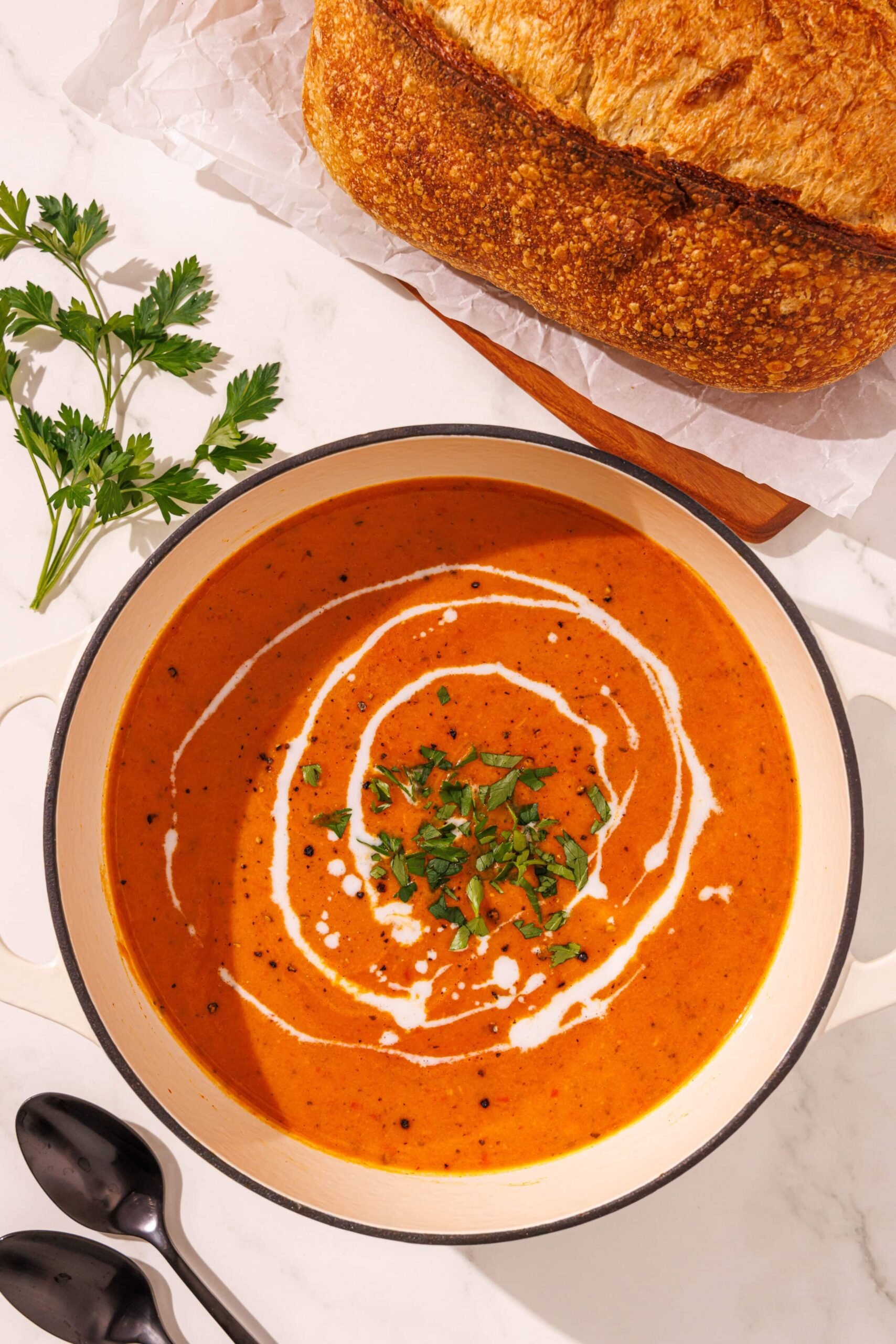 Glowing skin soup in a white ceramic bowl surrounded by two black spoons on the left, a sprig of parsley above, and a loaf of sourdough bread on a wood cutting board to the right.