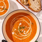 A large pot of red soup with a loaf of sliced bread beside the bowl, two metal black spoons, and a small bowl of soup.