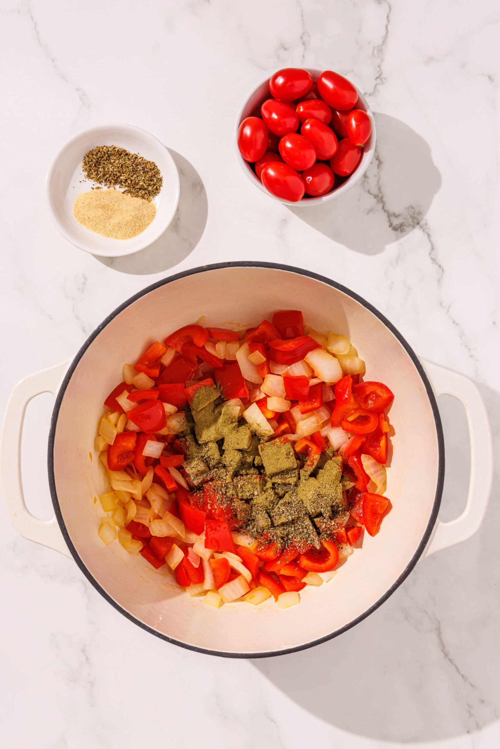 A white ceramic pot filled with bell peppers, onions, and spices. Above the bowl is a small white bowl of tomatoes and another small bowl of spices.