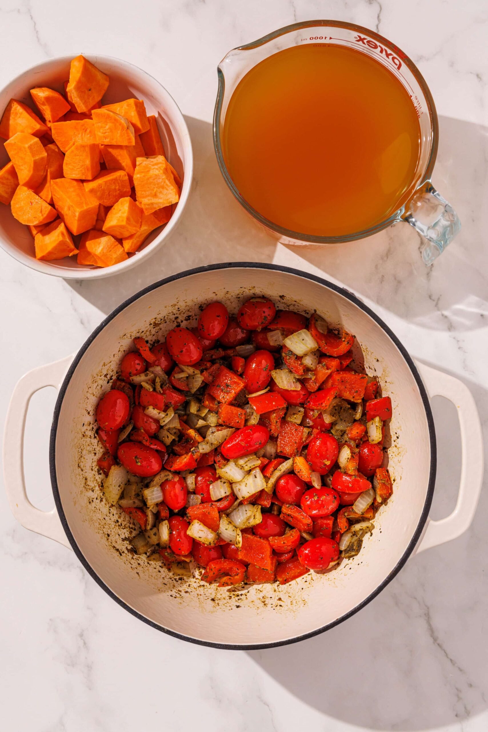 A white ceramic bowl filled bell pepper, onion, tomato, and spices. Above it is two other bowls with veggie broth and cubed sweet potato.