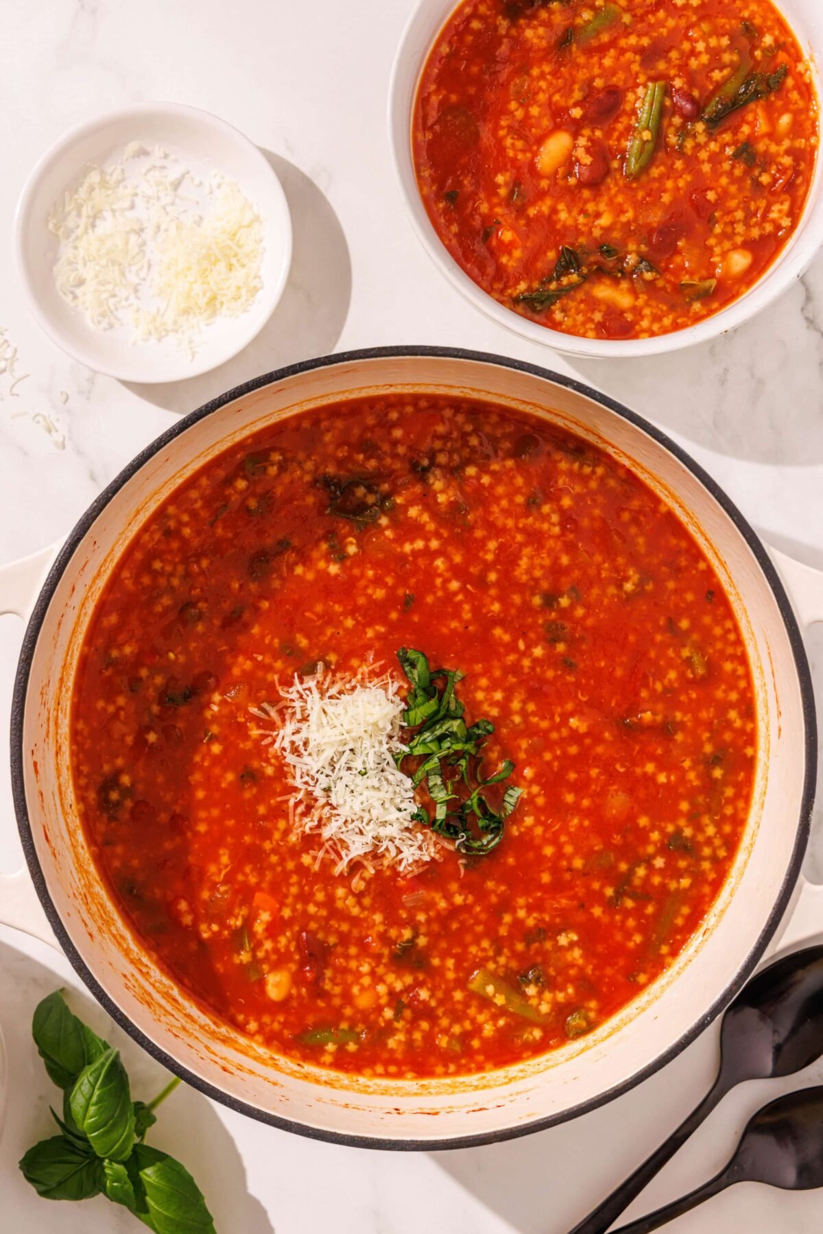 A large soup pot filled with a red soup topped with parmesan and basil. Beside the pot is a smaller bowl filled with soup, a small dish of parmesan, a basil leaf, and two black metal spoons.