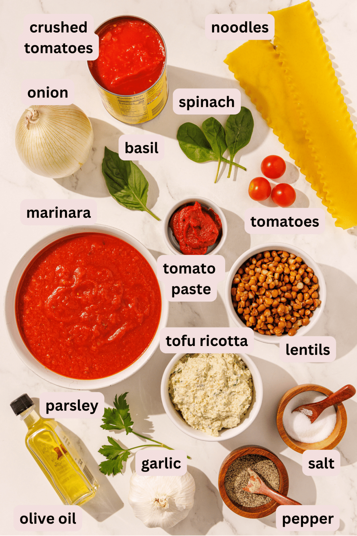 Ingredients laid out on a counter and labeled including: crushed tomatoes, noodles, spinach, onion, basil, cherry tomatoes, marinara, tomato paste, lentils, tofu ricotta, salt, papper, garlic, parsley, and olive oil.