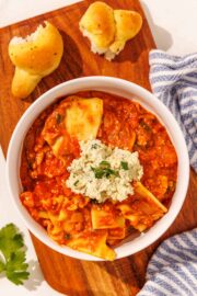 A white bowl of lasagna soup topped with a scoop of ricotta. The bowl is sitting on a brown cutting board with two garlic knots on it and a blue and white striped towel as well.