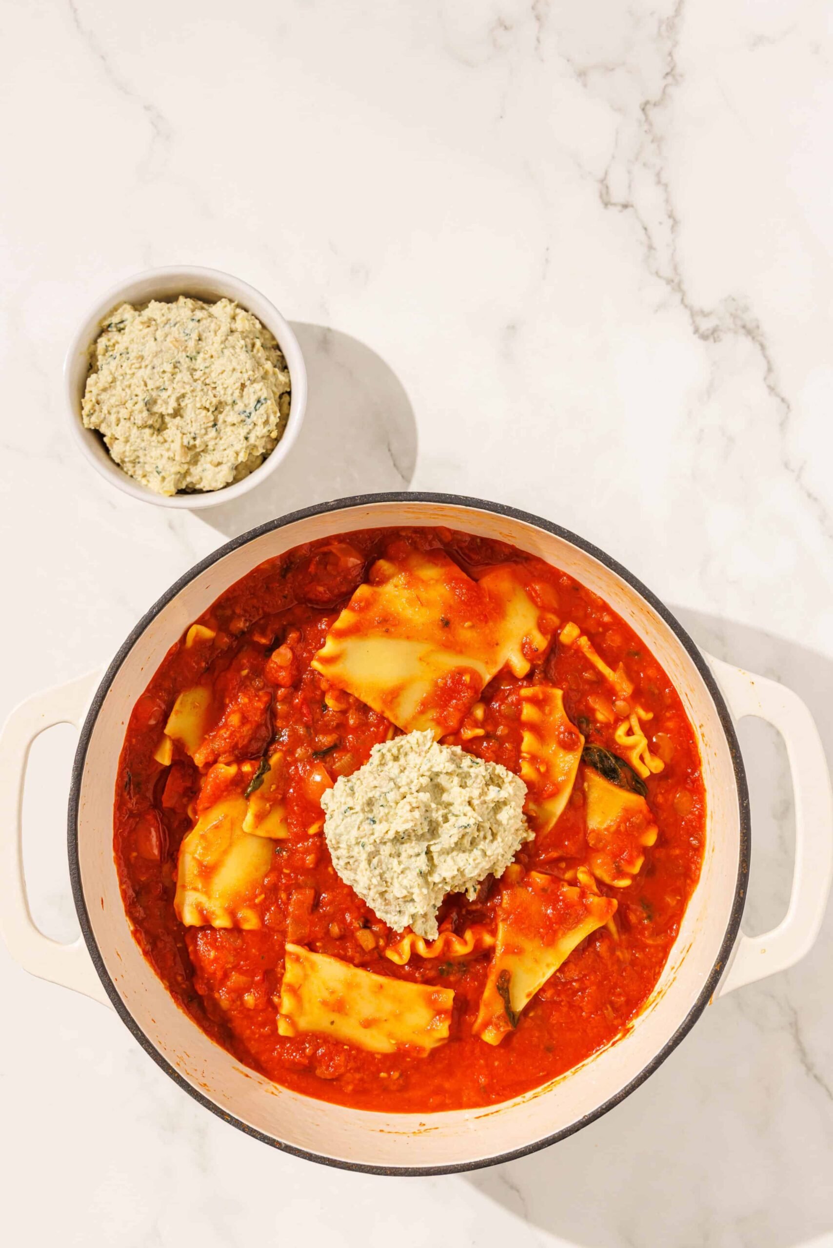 A large soup pot filled with lasagna soup with a dollop of ricotta in the center. To the left of the bowl is a smaller bowl of ricotta.